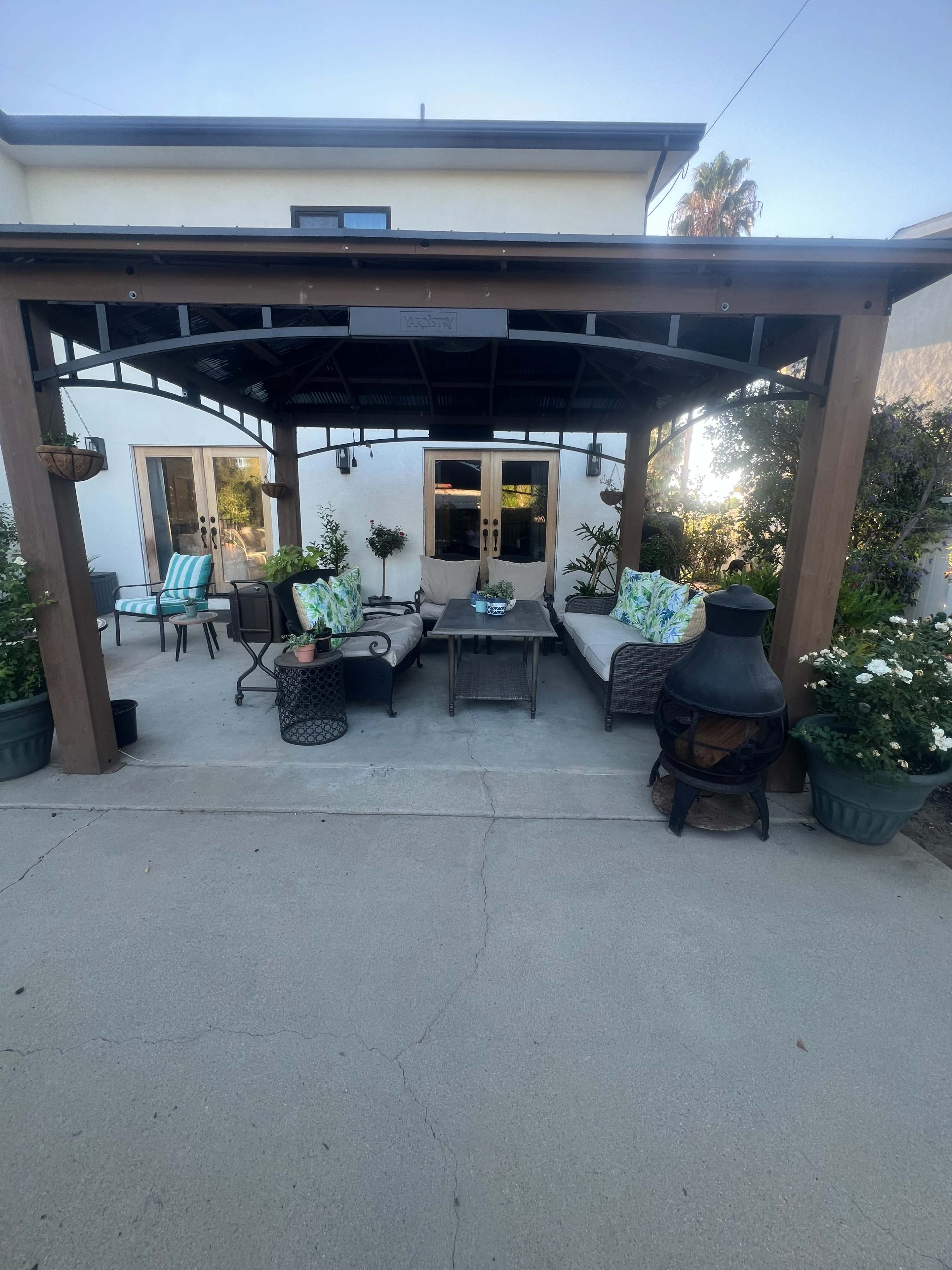 The image shows a covered outdoor seating area with comfortable furniture, including sofas and chairs, surrounding a small table, and a chimney-style heater nearby, in a well-maintained backyard.