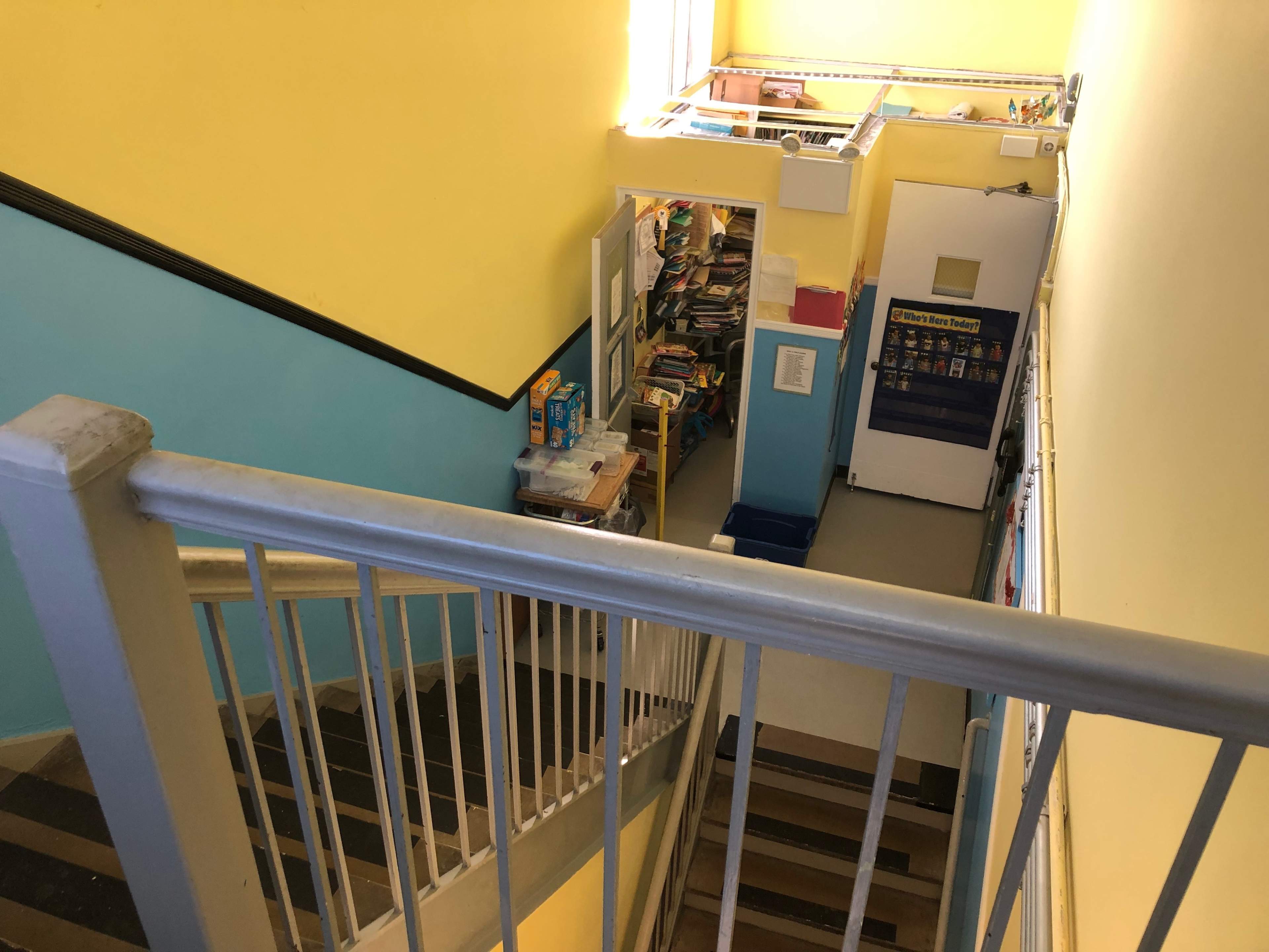 A staircase leads down to a brightly colored hallway with shelves of books and supplies on the right.