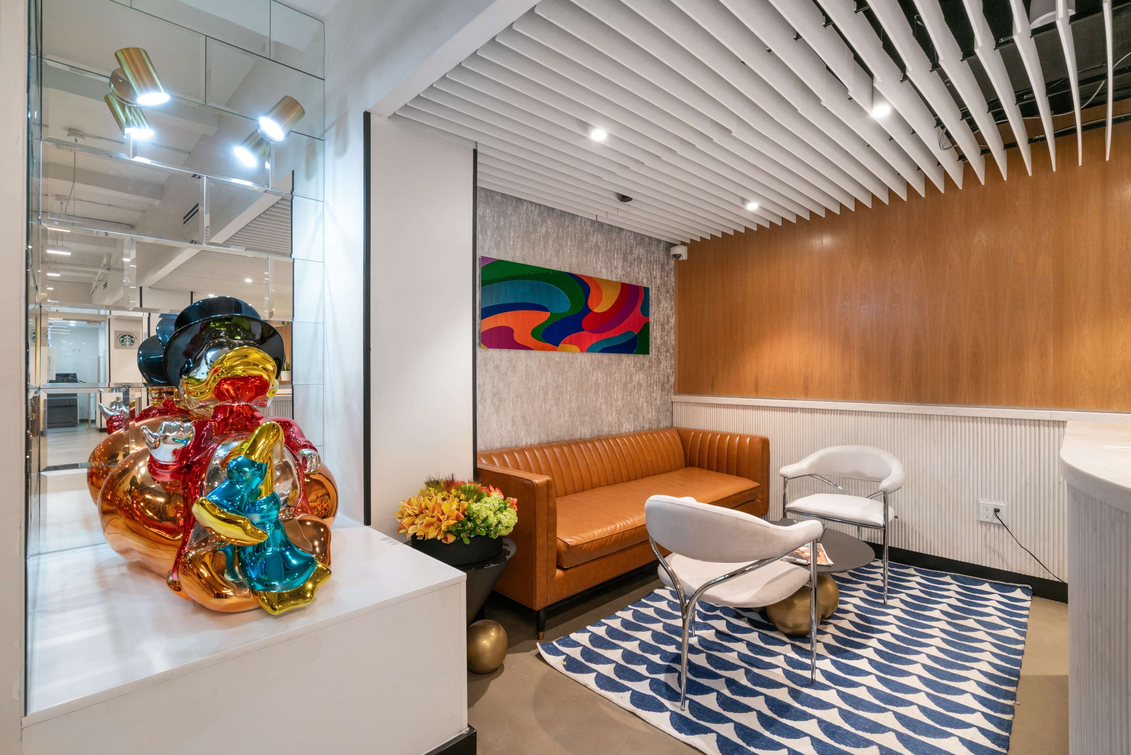 The image displays a modern reception area featuring an orange sofa, a white chair, colorful abstract wall art, and a glossy sculptural piece alongside a small arrangement of flowers.