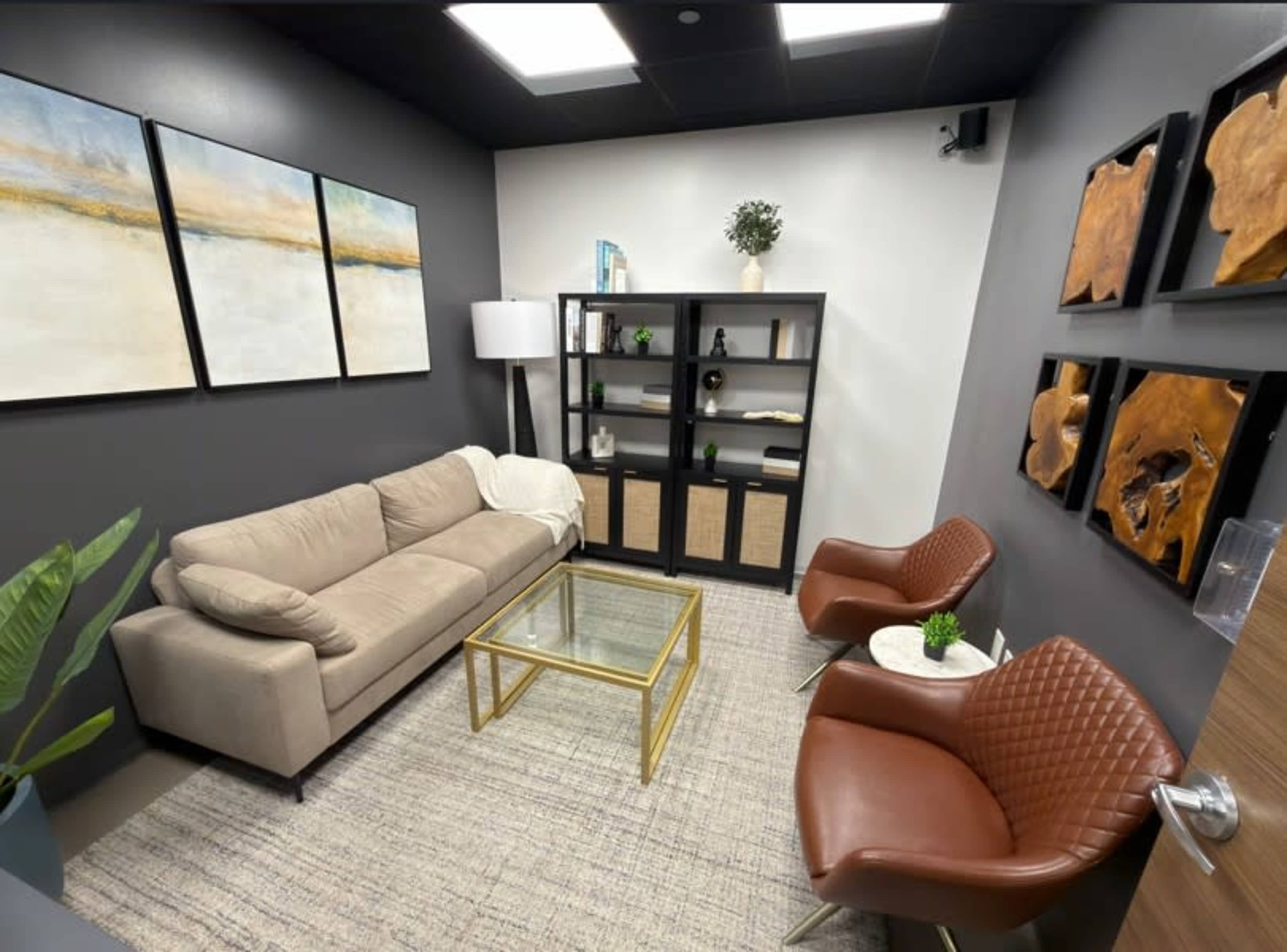 The image shows a small, modern waiting room featuring a beige sofa, two brown chairs, a glass coffee table, and a black shelf with decorative items, all set against gray walls.