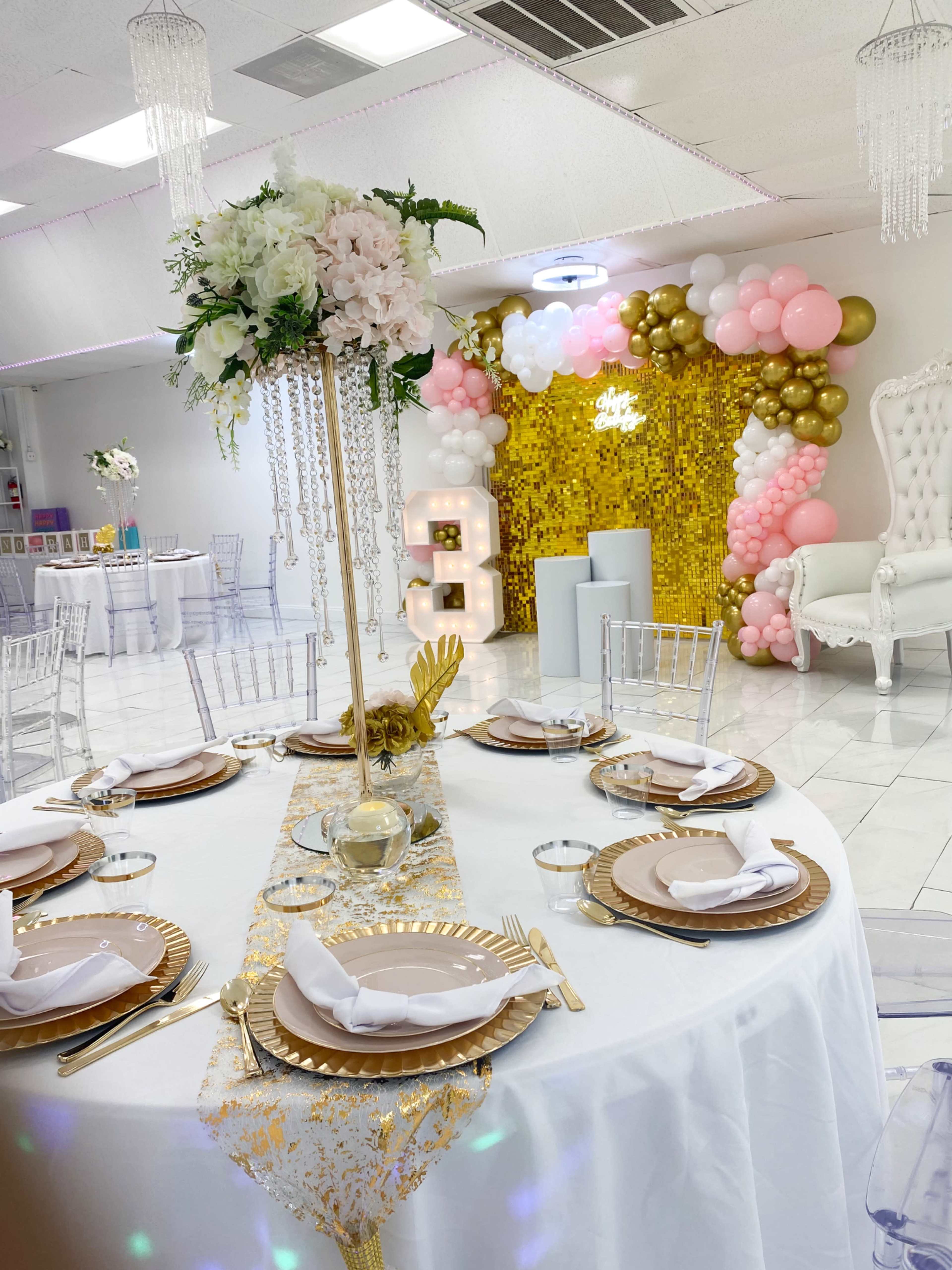 A beautifully arranged banquet table with gold chargers, white napkins, and a tall floral centerpiece, set against a backdrop of pastel balloons and a sparkling golden wall.
