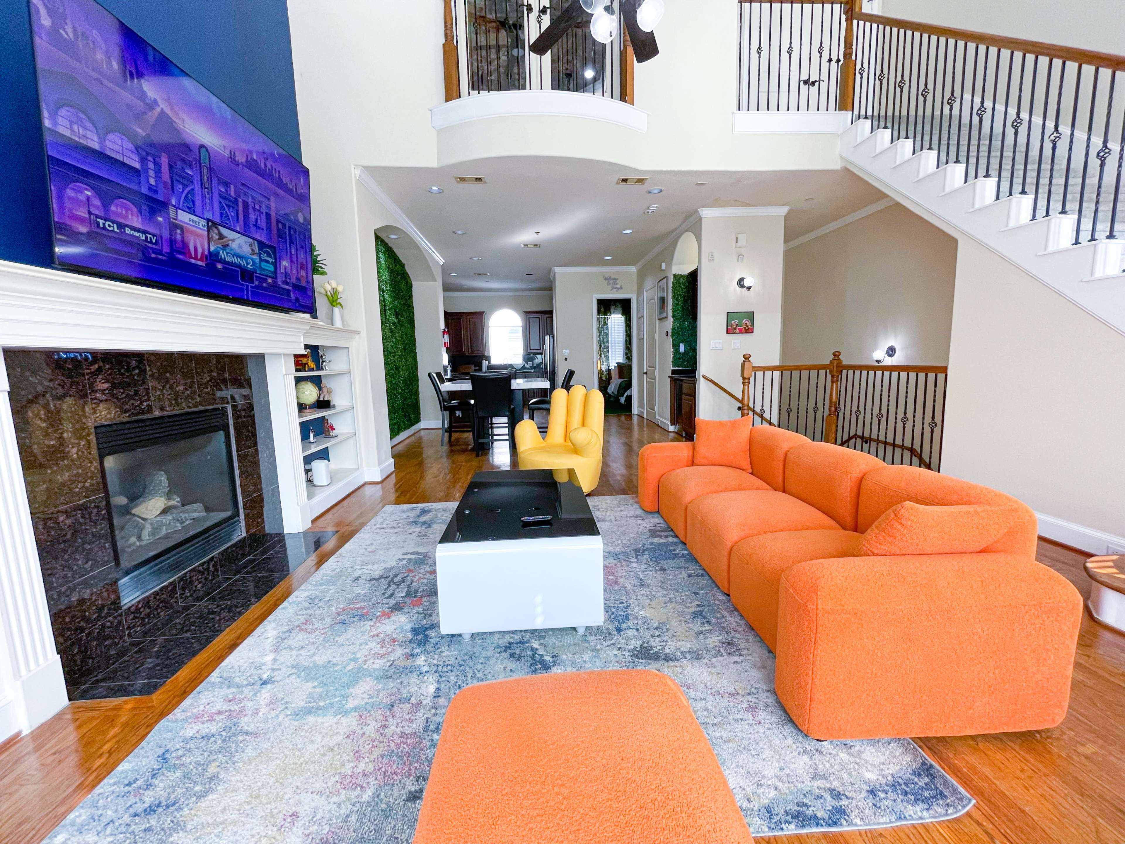 A spacious living room features a large orange sectional sofa, a yellow chair, a modern coffee table, and a television mounted above a fireplace.