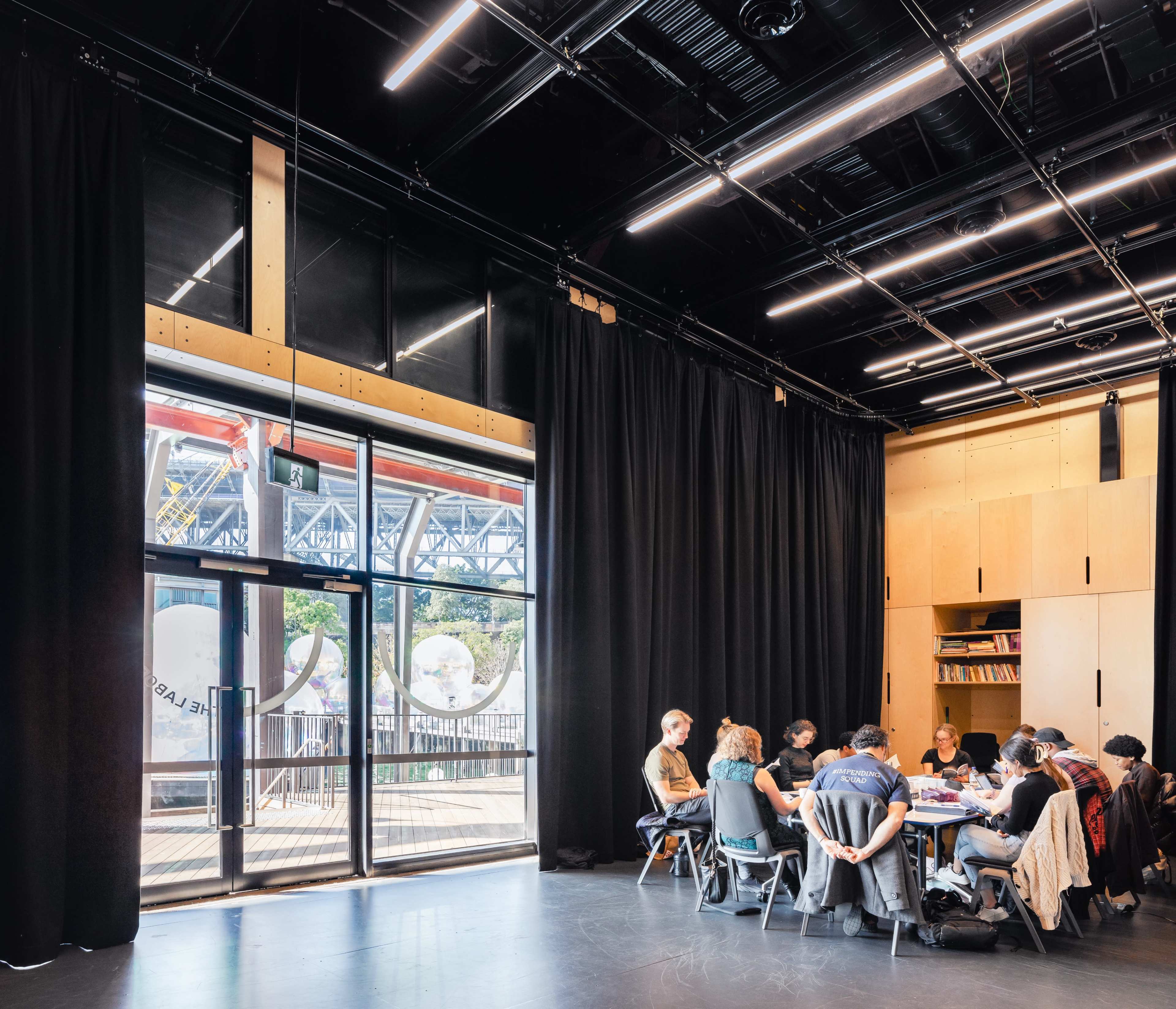 A group of people sits around a table in a well-lit room with large windows and black curtains, overlooking an industrial landscape.