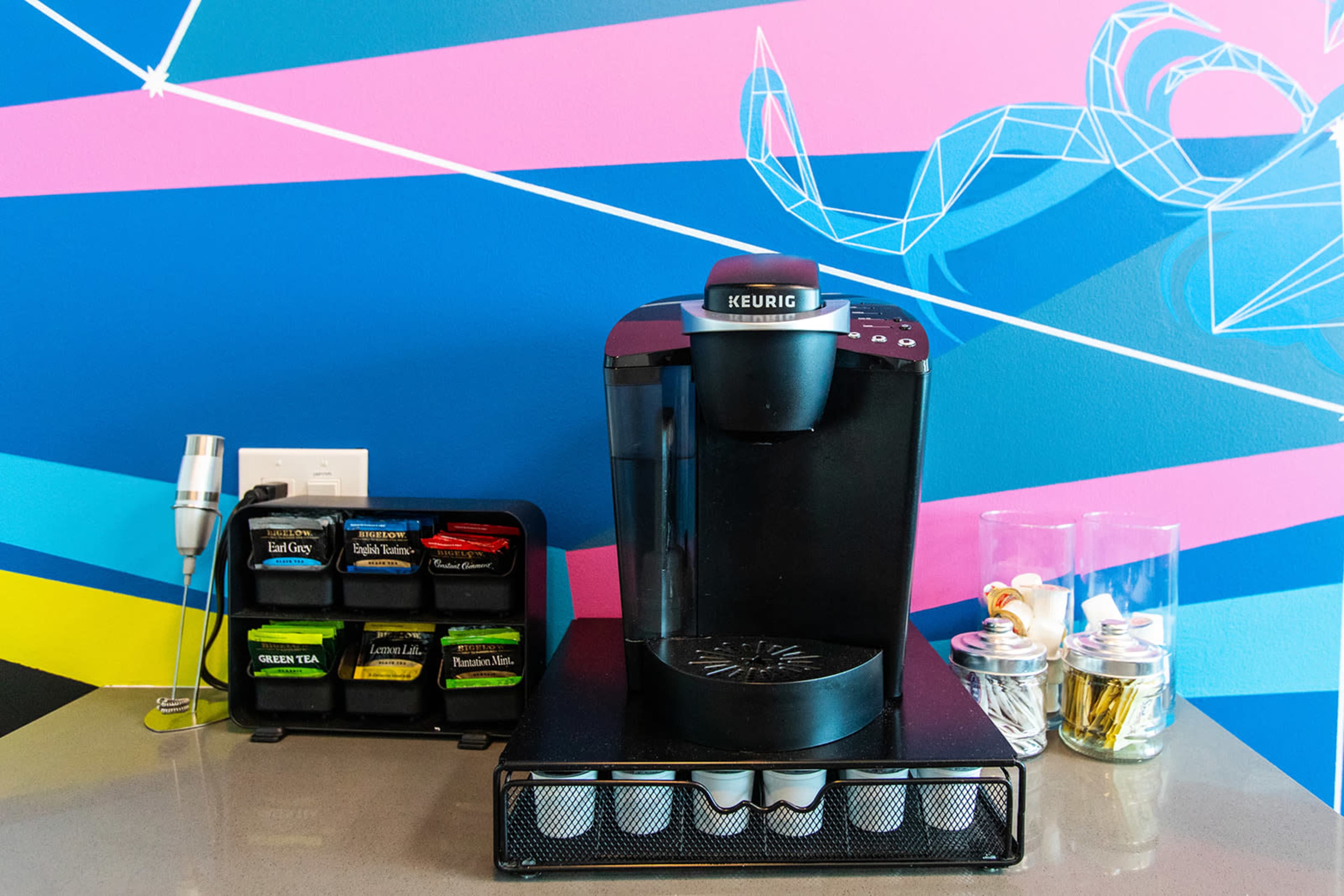 A Keurig coffee machine is placed on a countertop next to a variety of coffee and tea pods, against a colorful geometric wall.