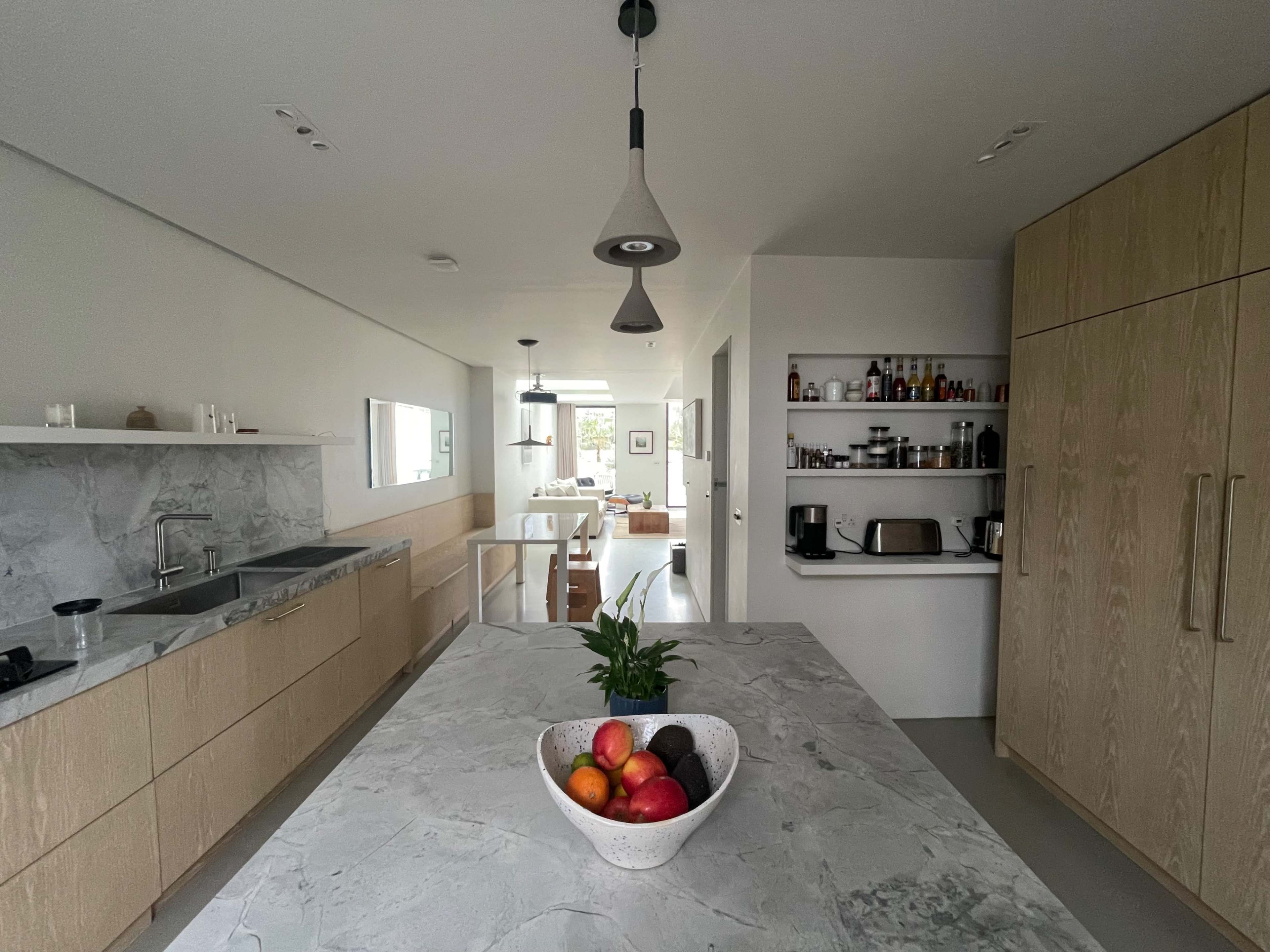 The image shows a modern kitchen with a large marble island, light wood cabinetry, and a bowl of colorful fruit on the island.