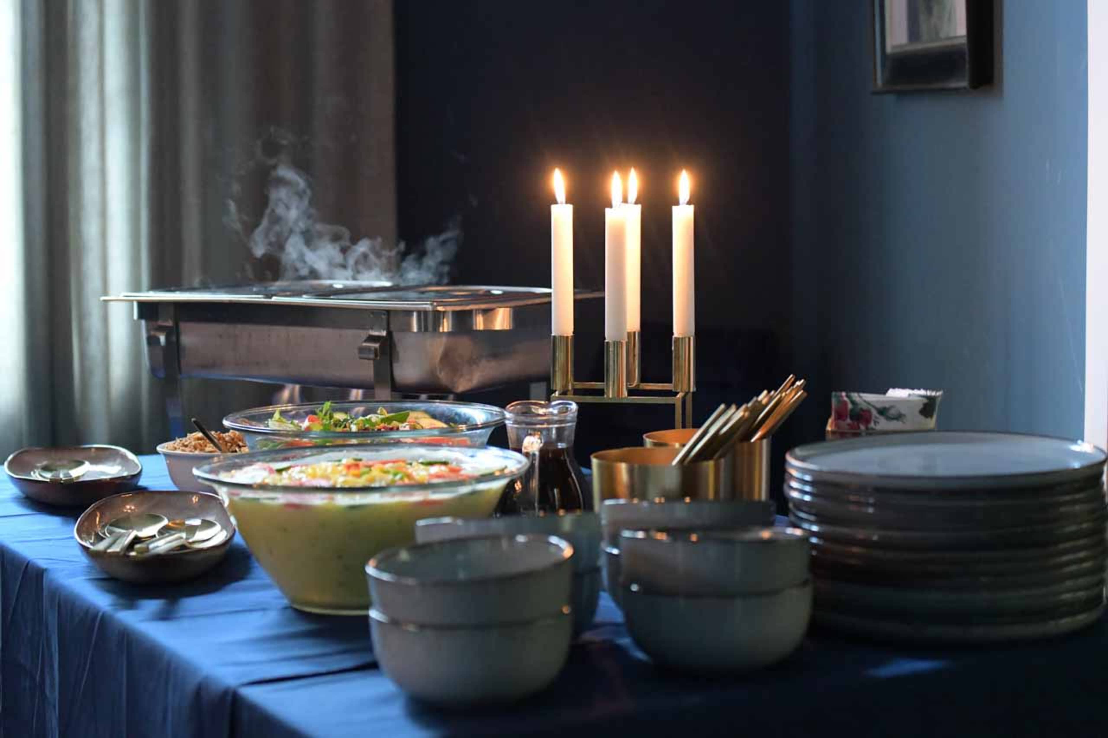 A table is set with a variety of food in glass bowls, candles lit, and stacked plates in a softly lit room.