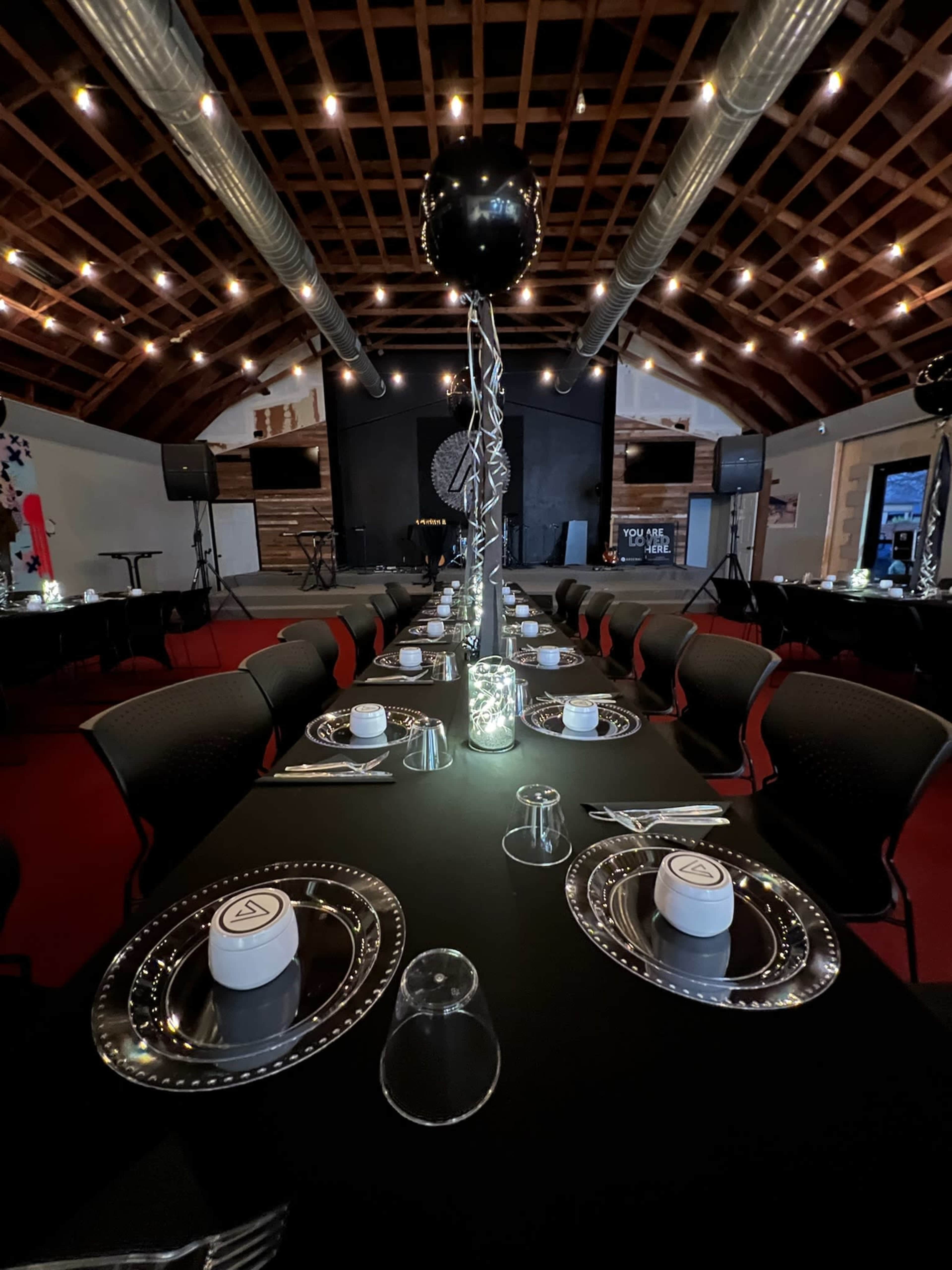 The image shows a long dining table set with plates, cups, and a decorative centerpiece in a venue with wooden beams and overhead lighting.