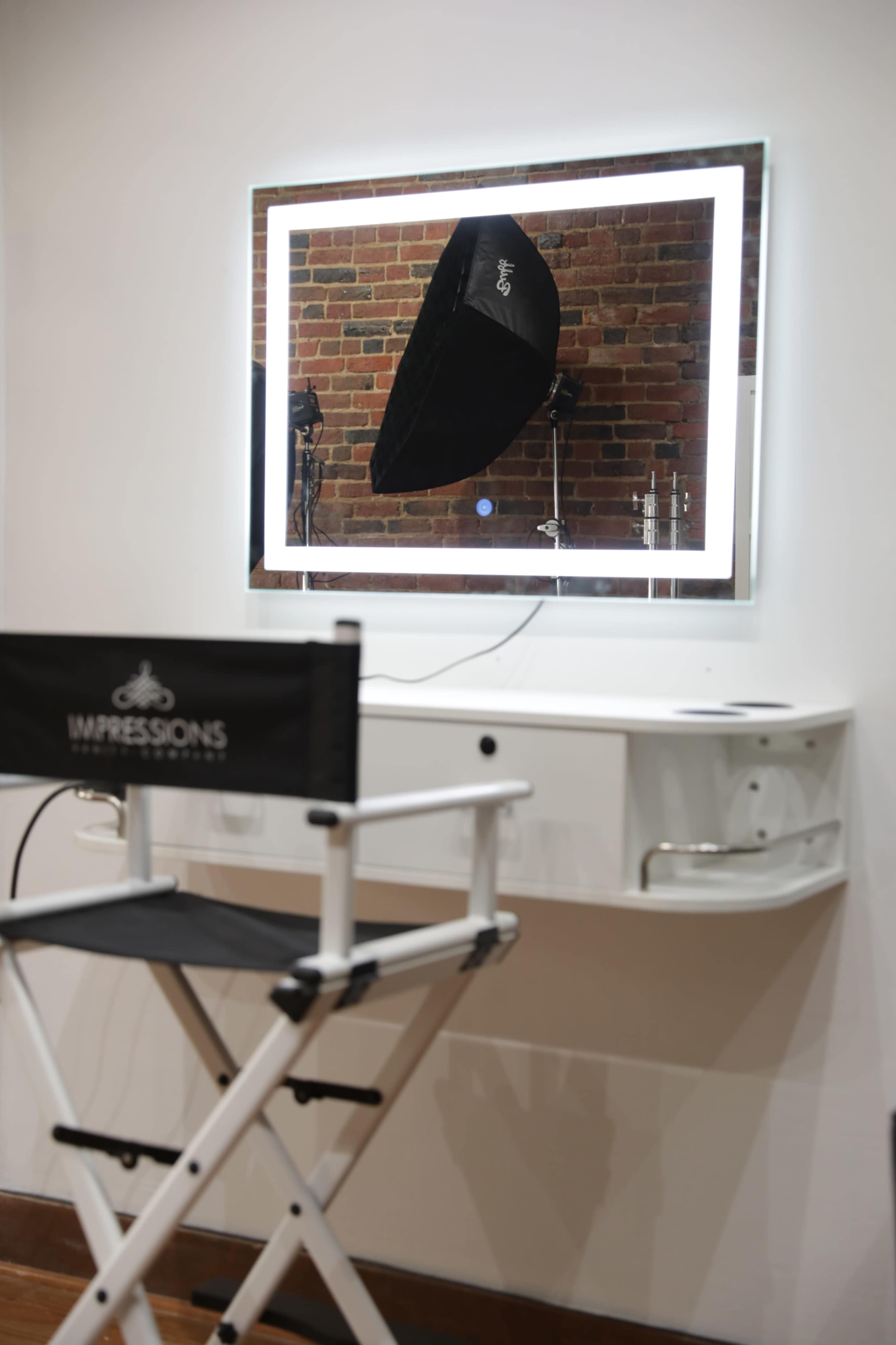 A makeup chair is positioned in front of a backlit mirror in a room with a brick wall.