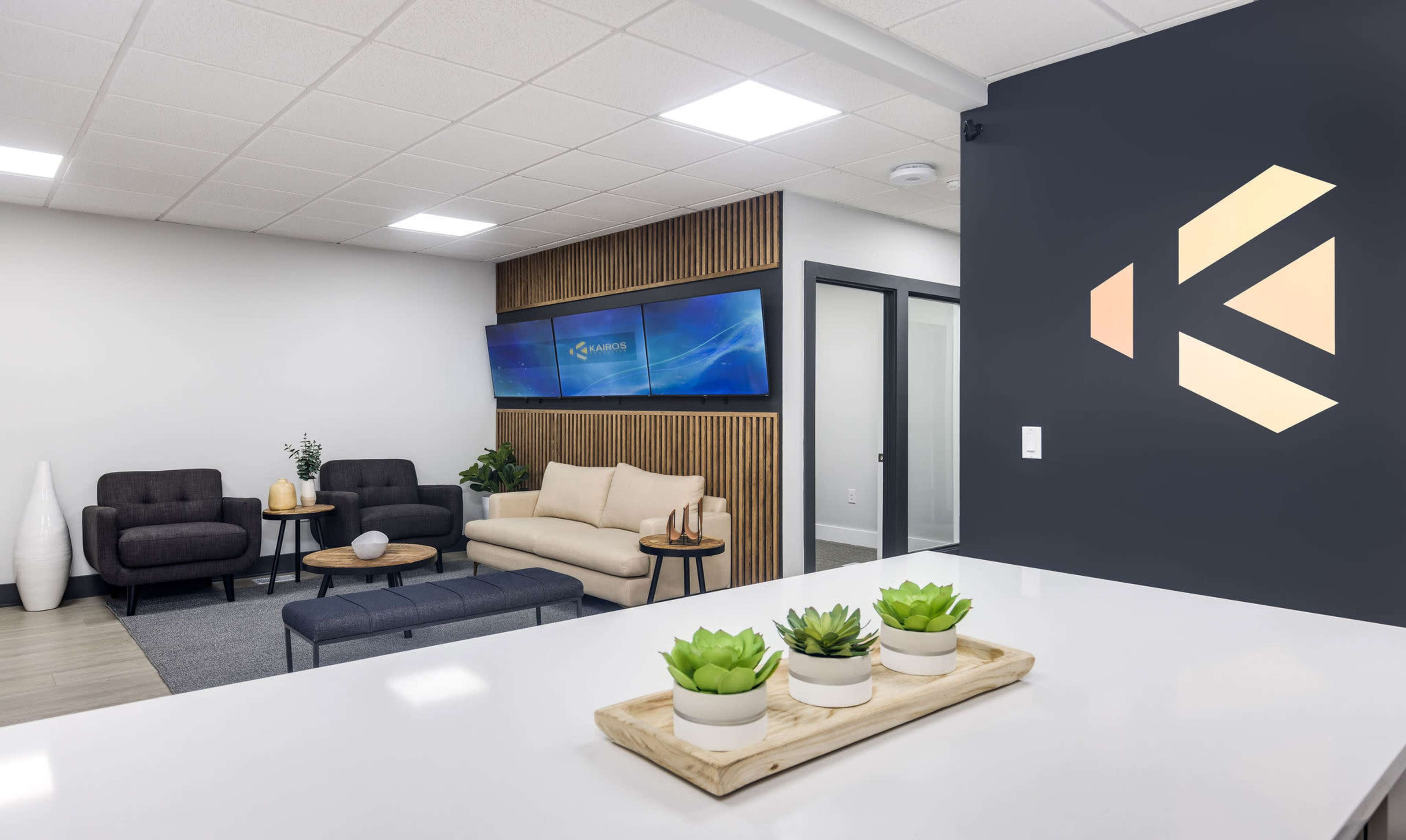 A modern office reception area features a seating arrangement with two sofas, a wall-mounted television, and decorative plants on a counter.