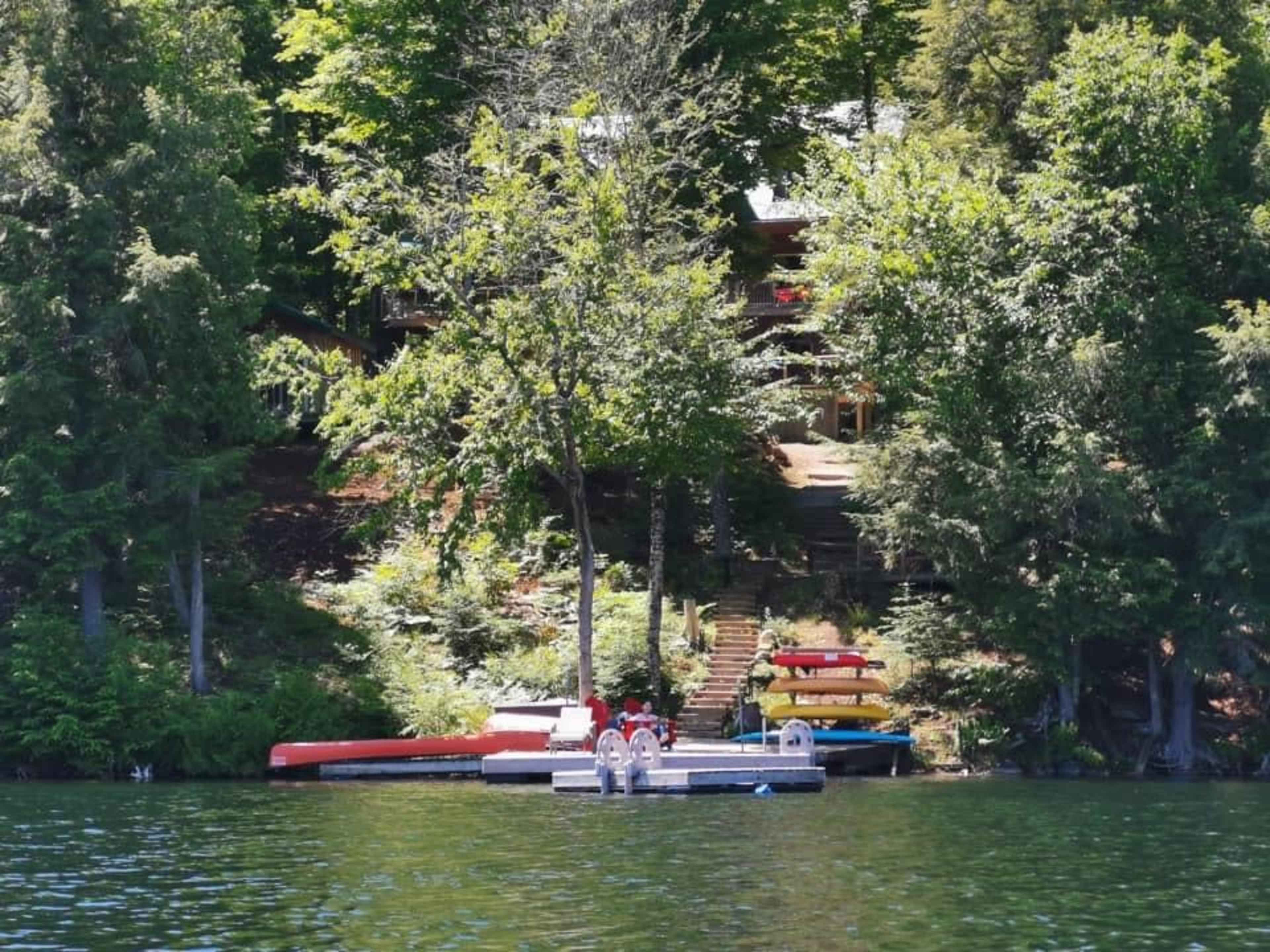 A dock with several boats is situated in front of a forested area, leading up to a staircase and a cabin among the trees.