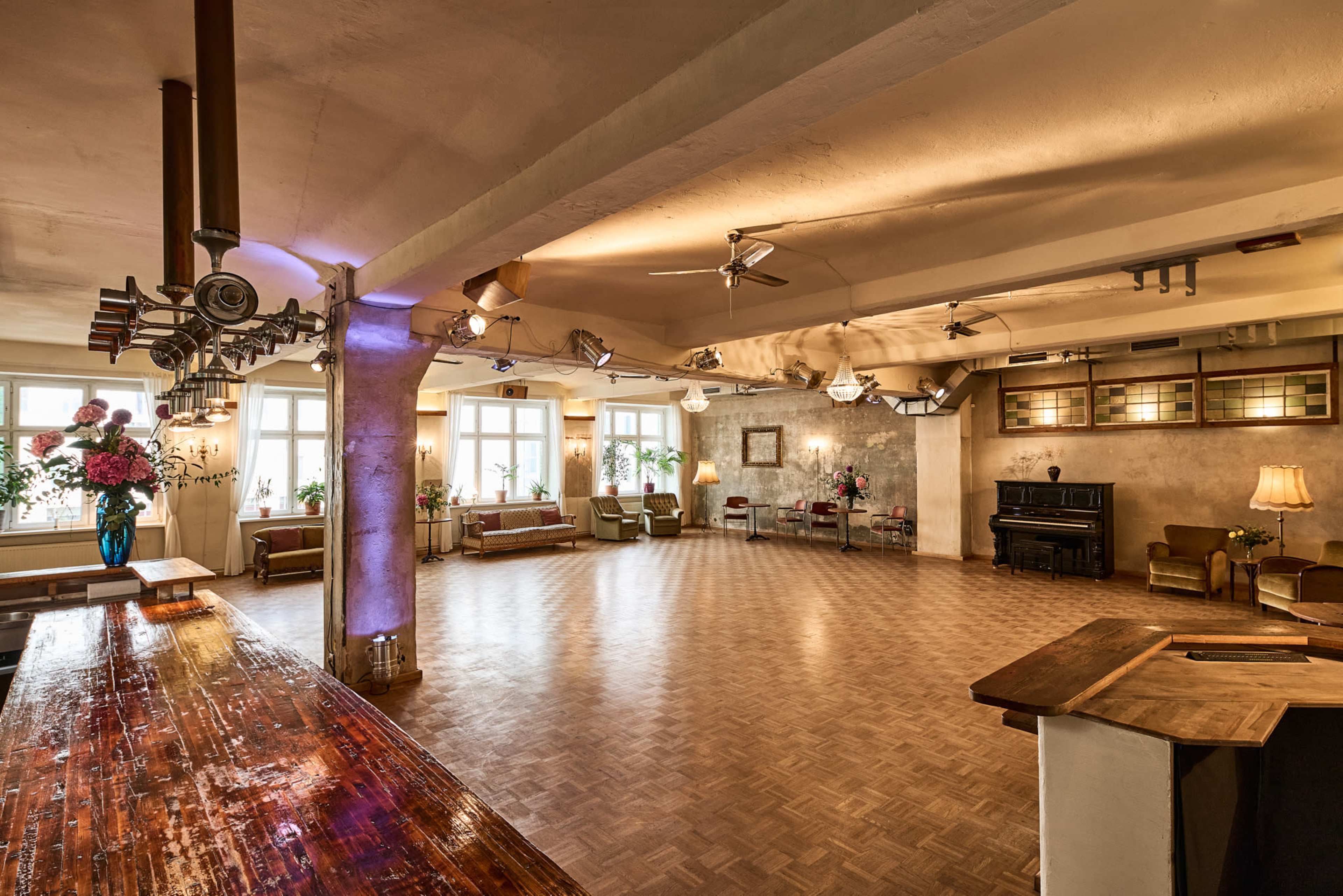 A spacious, well-lit room features wooden floors, a bar area, several seating arrangements, and large windows with plants.