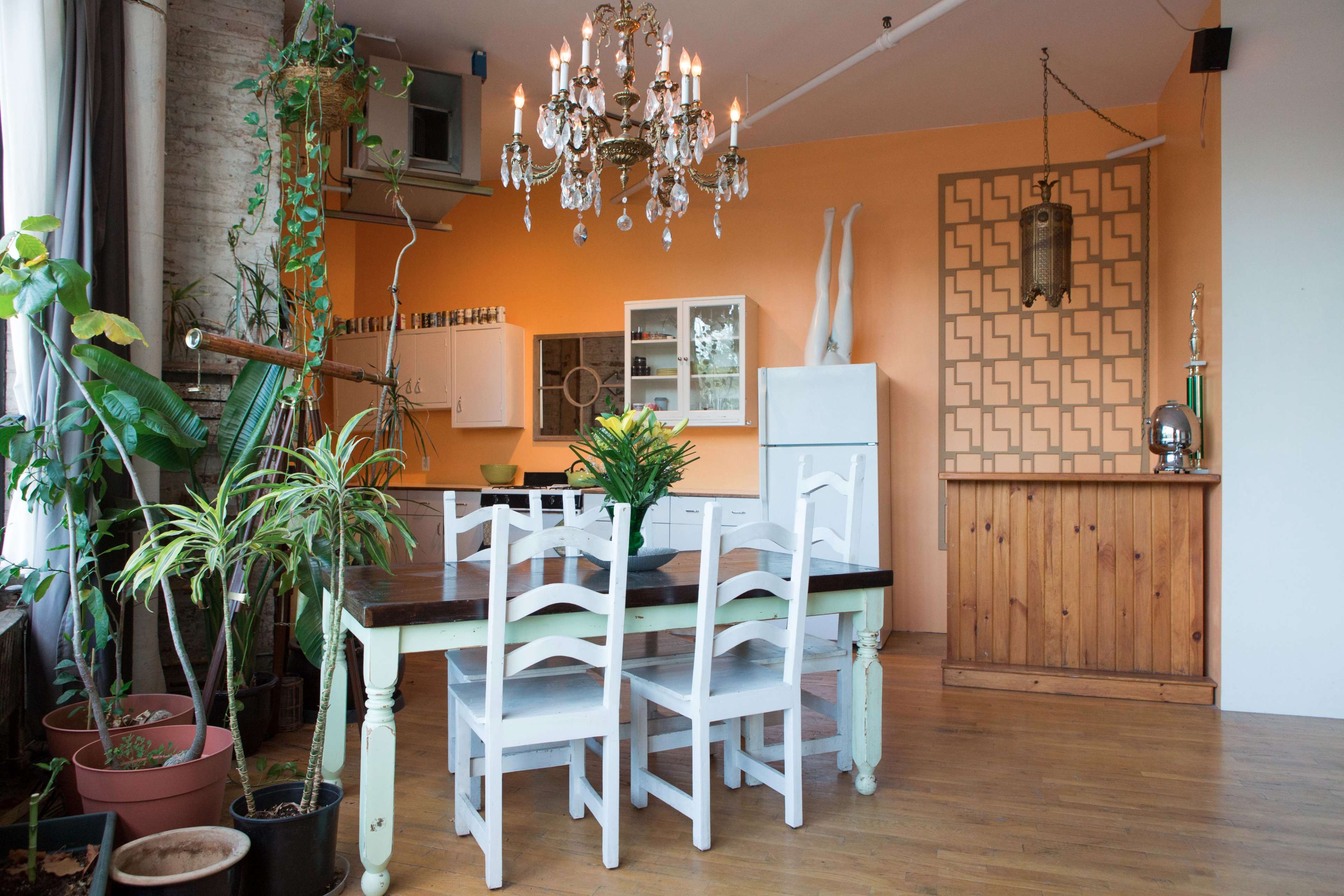A dining area features a wooden table surrounded by white chairs, a chandelier hangs from the ceiling, and a backdrop of orange walls with decorative patterns.