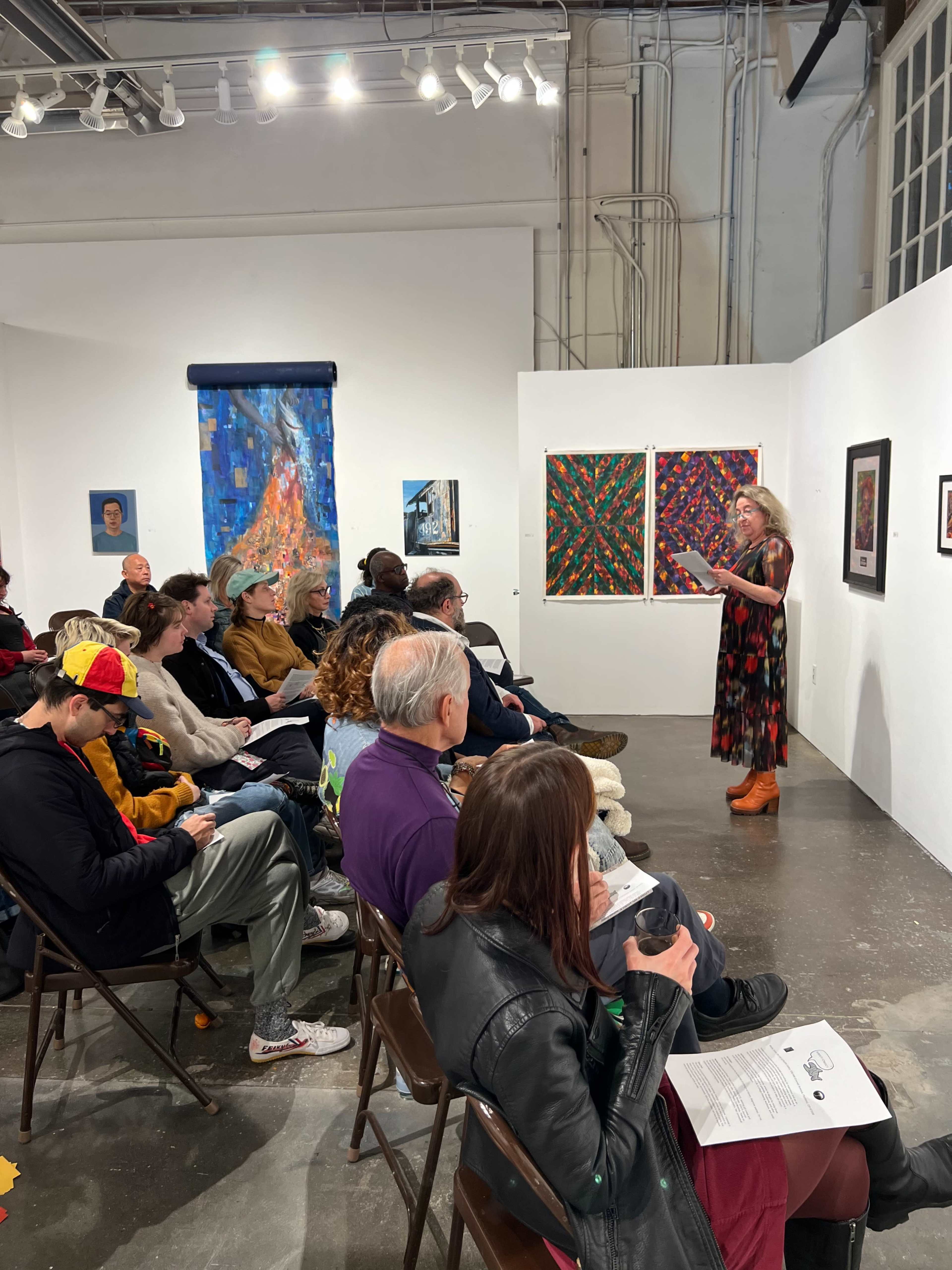A woman in a dress stands at the front of an art gallery, reading to an audience seated in folding chairs, surrounded by colorful artwork on the walls.