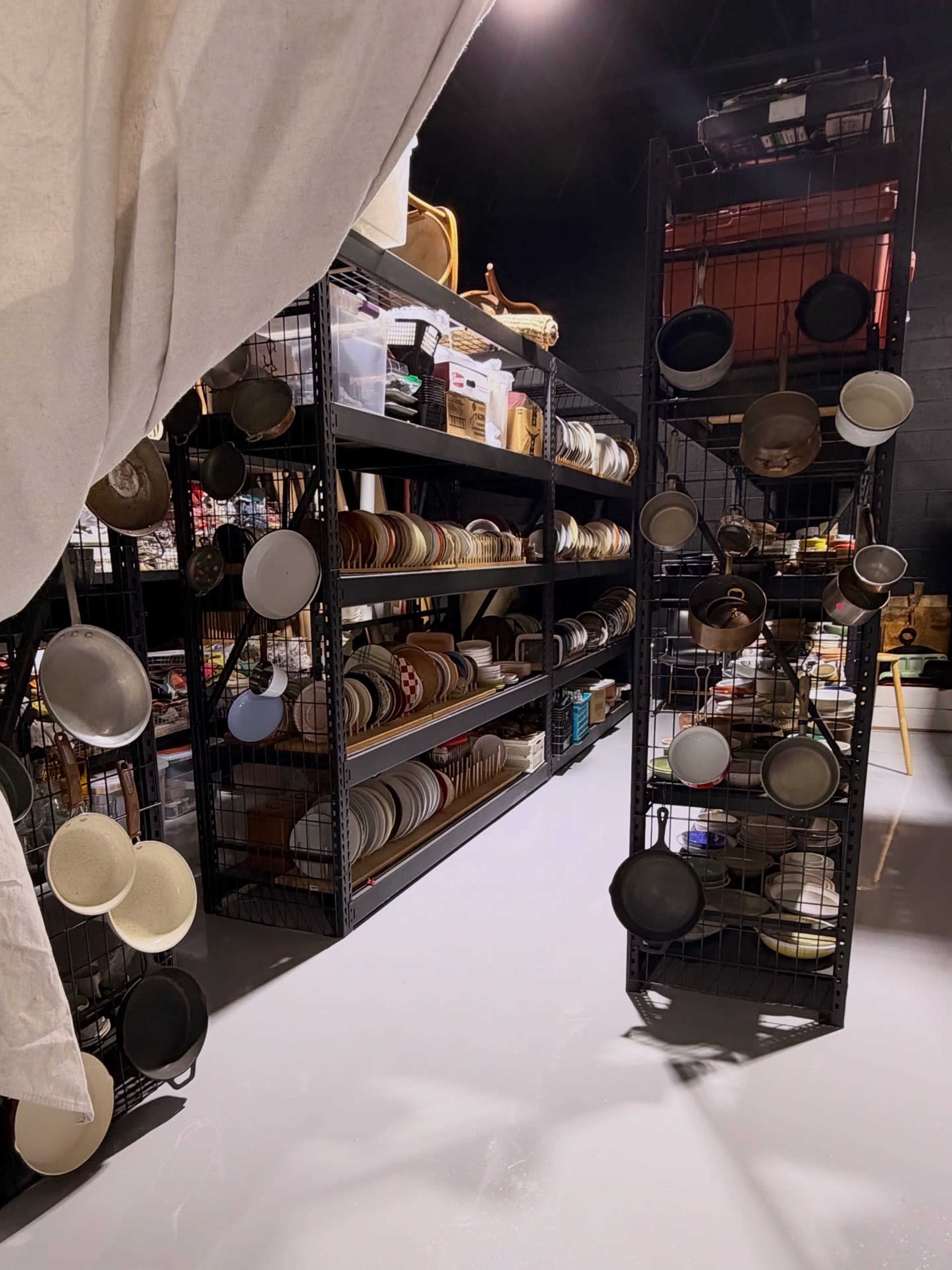 The image shows a storage area with metal shelves filled with various types of dishware, while cookware hangs from a grid rack in the foreground.