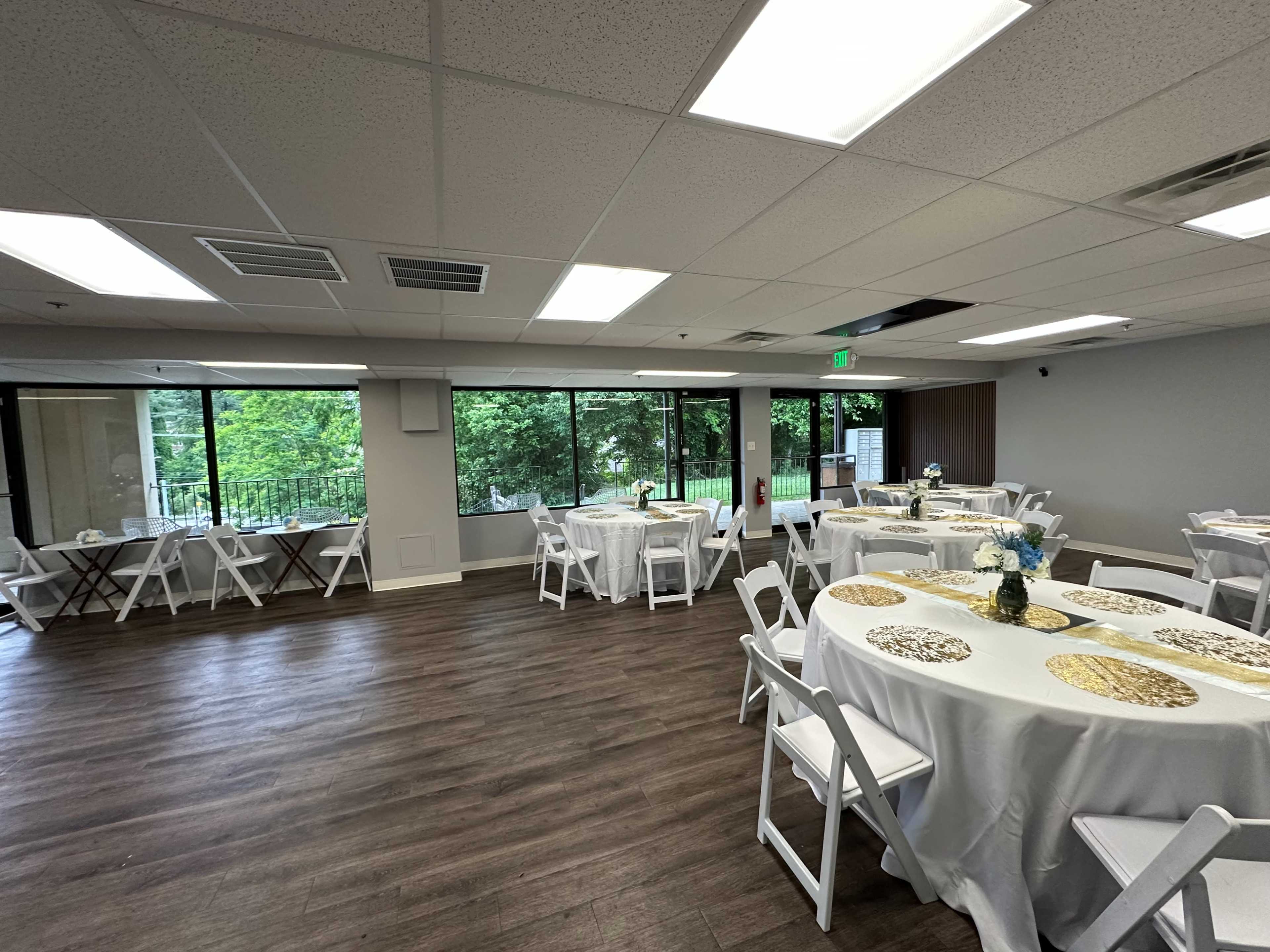 The image shows a banquet room with round tables covered in white tablecloths, each adorned with decorative centerpieces, and large windows overlooking greenery.