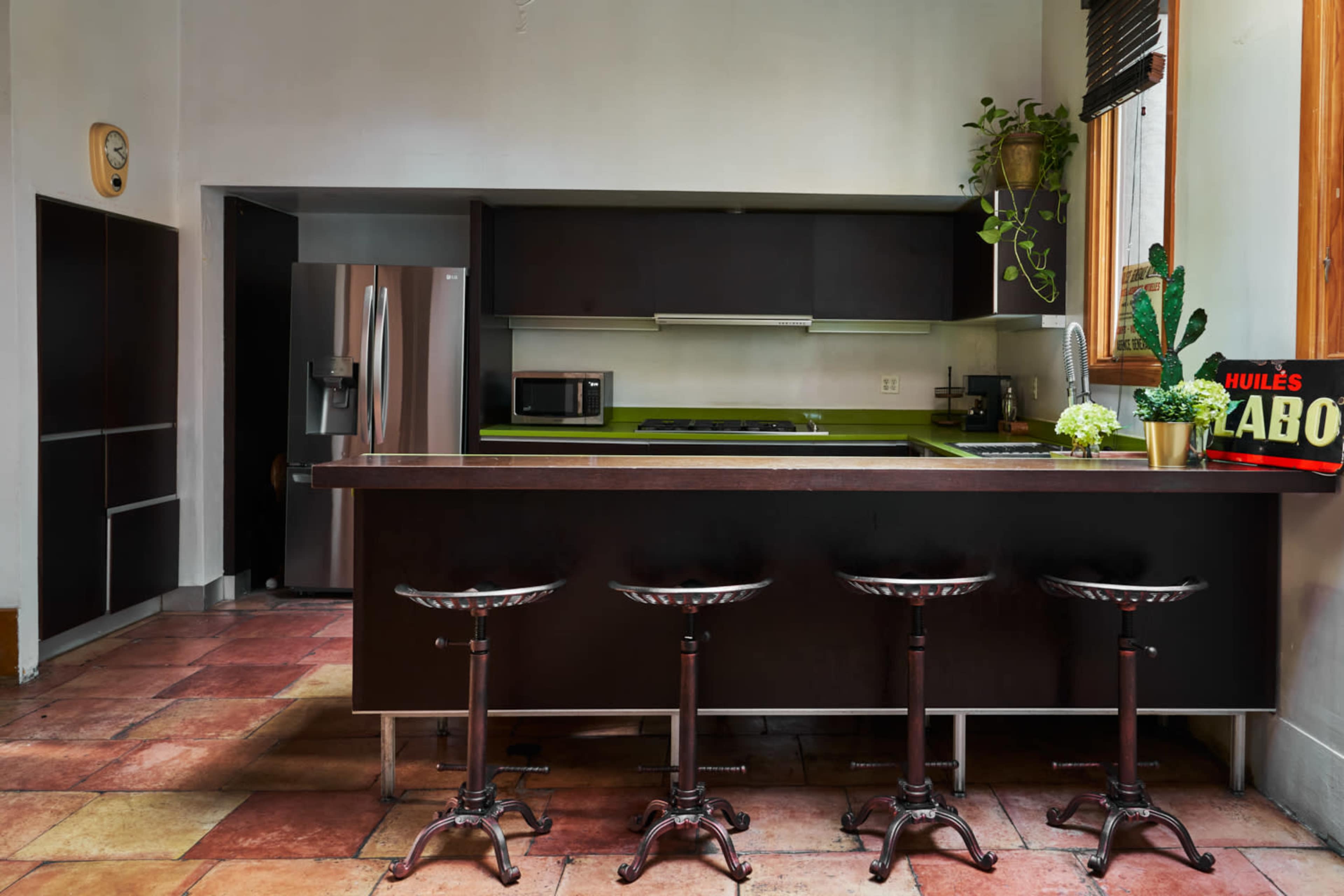 A modern kitchen features a central island with four metal stools, dark cabinetry, and a stainless steel refrigerator against the wall.