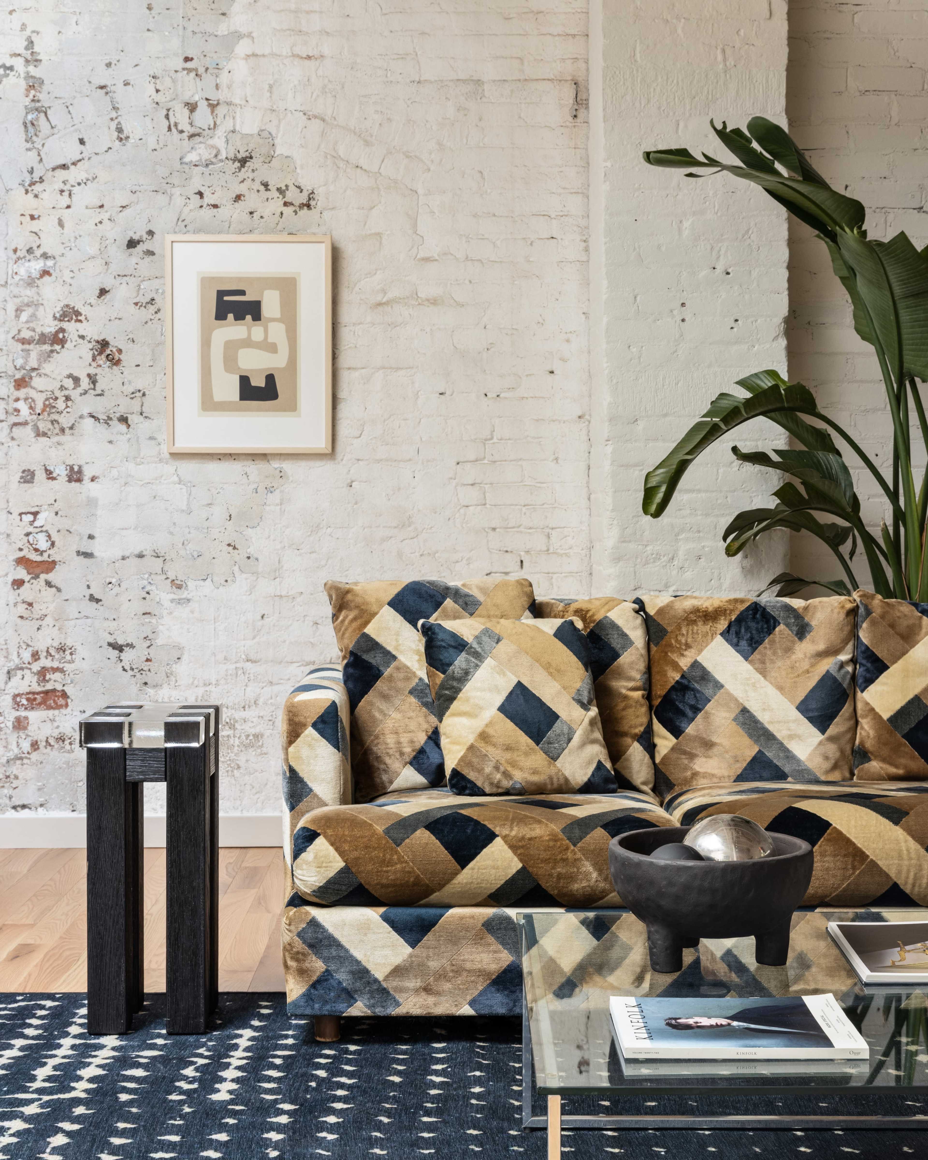 A modern living room featuring a patterned sofa, a glass coffee table, a decorative bowl, and a tall plant against a white brick wall.