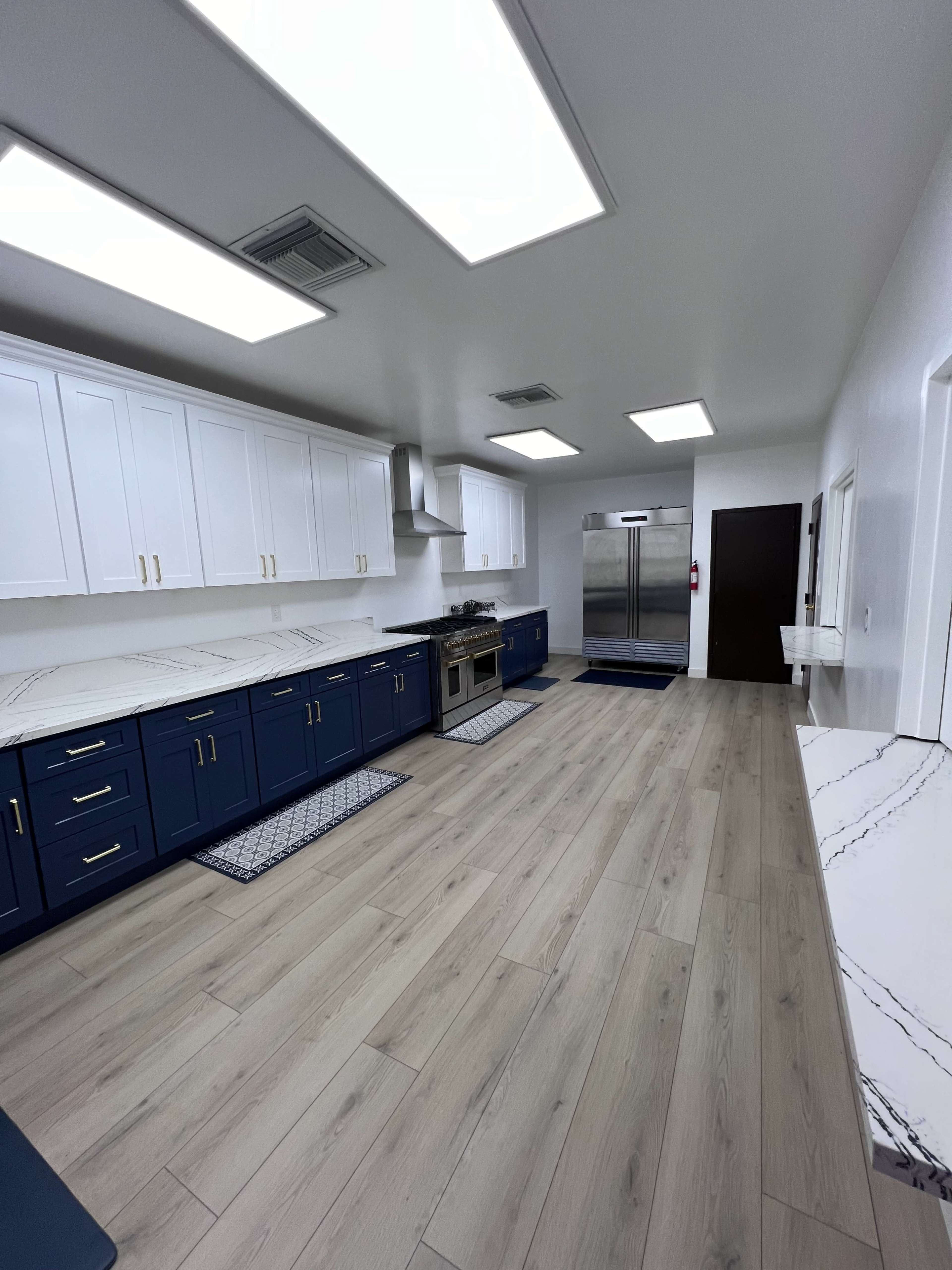 The image shows a modern kitchen with white upper cabinets, blue lower cabinets, a large stainless steel refrigerator, and white marble countertops, all set against light wooden flooring.