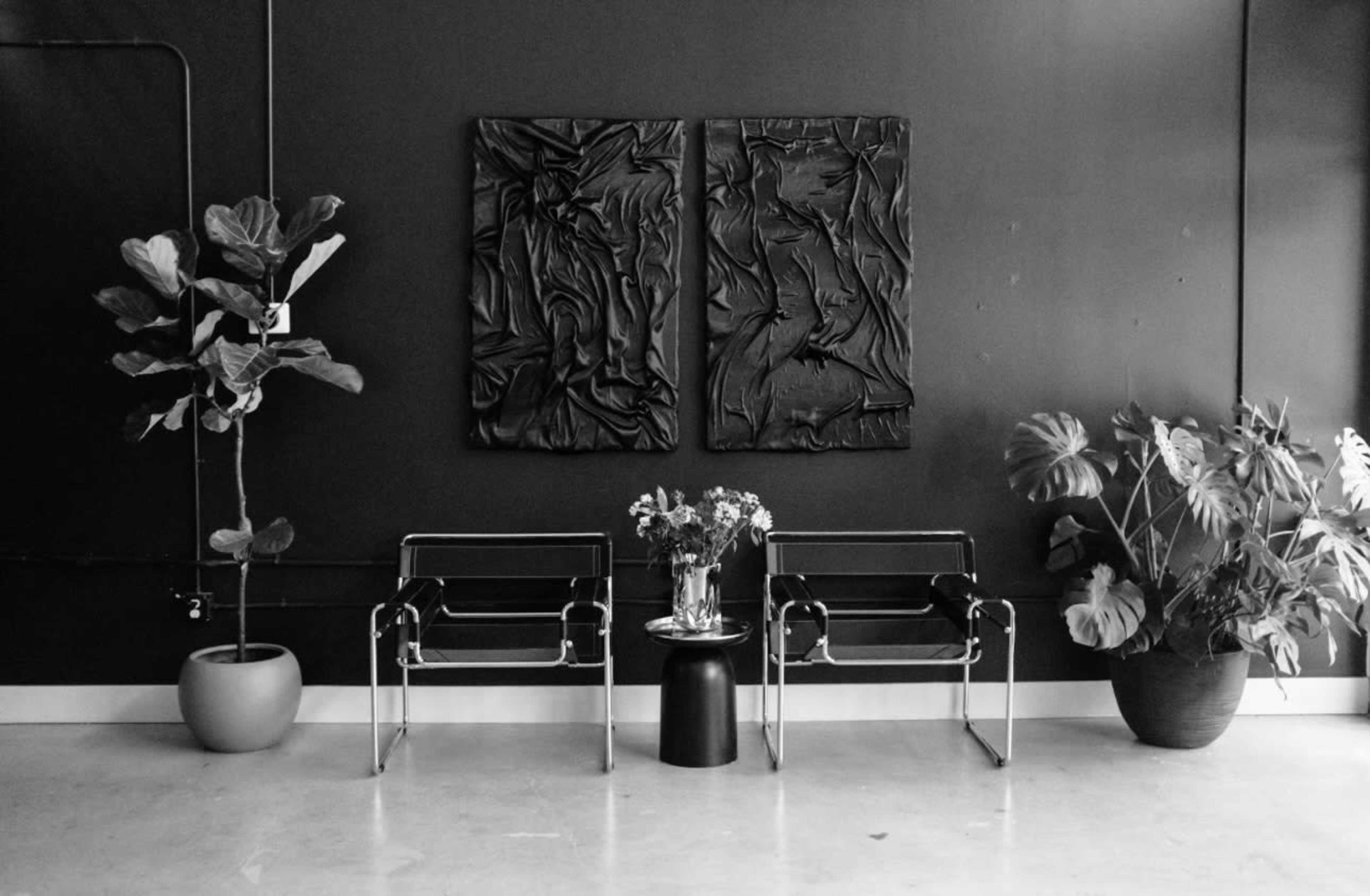 Two modern chairs flanking a small table with a vase of flowers, against a backdrop of abstract artwork and potted plants.