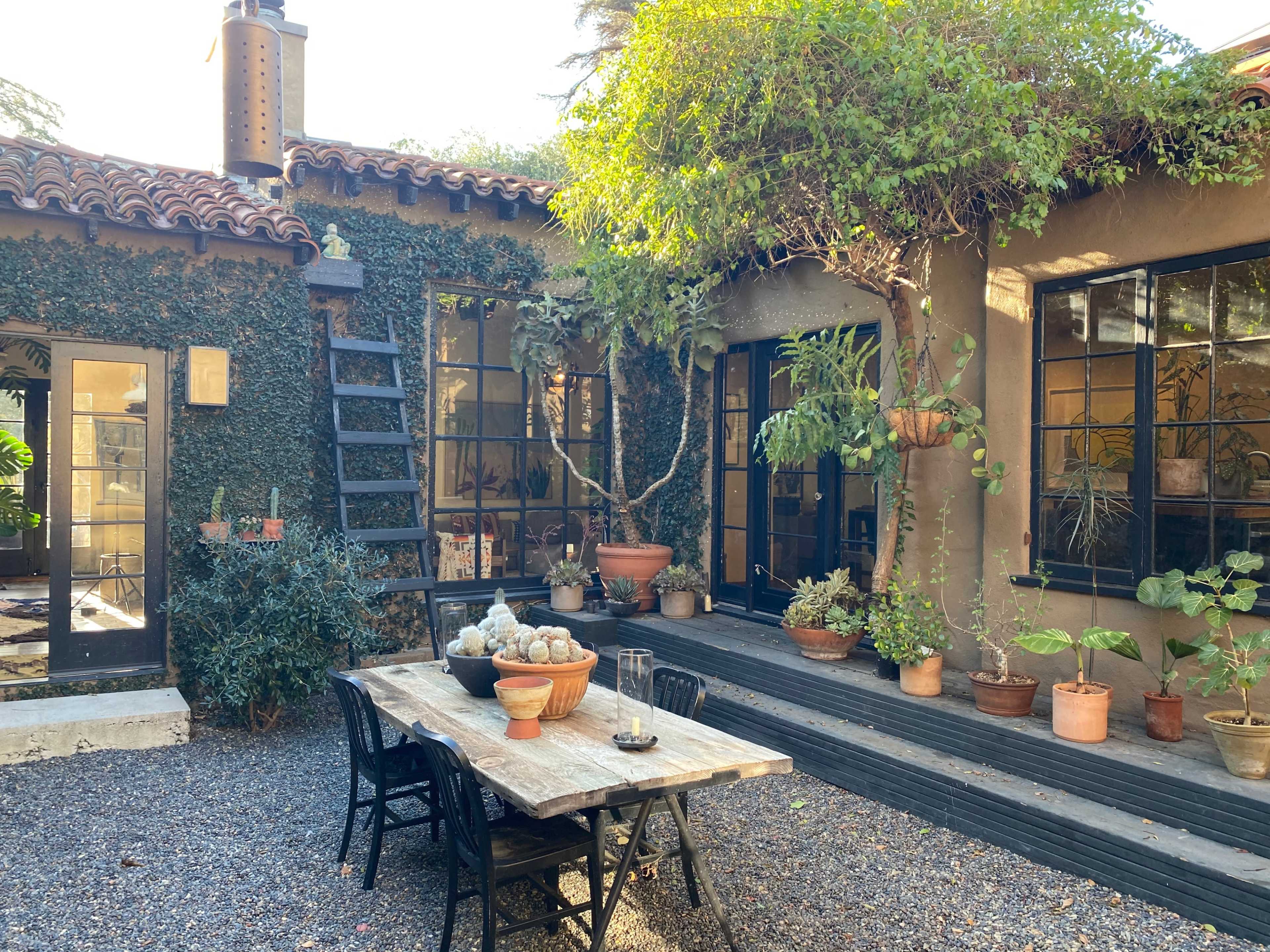 A rustic outdoor courtyard features a wooden table surrounded by potted plants and a stone pathway, with a vine-covered wall and large windows in the background.