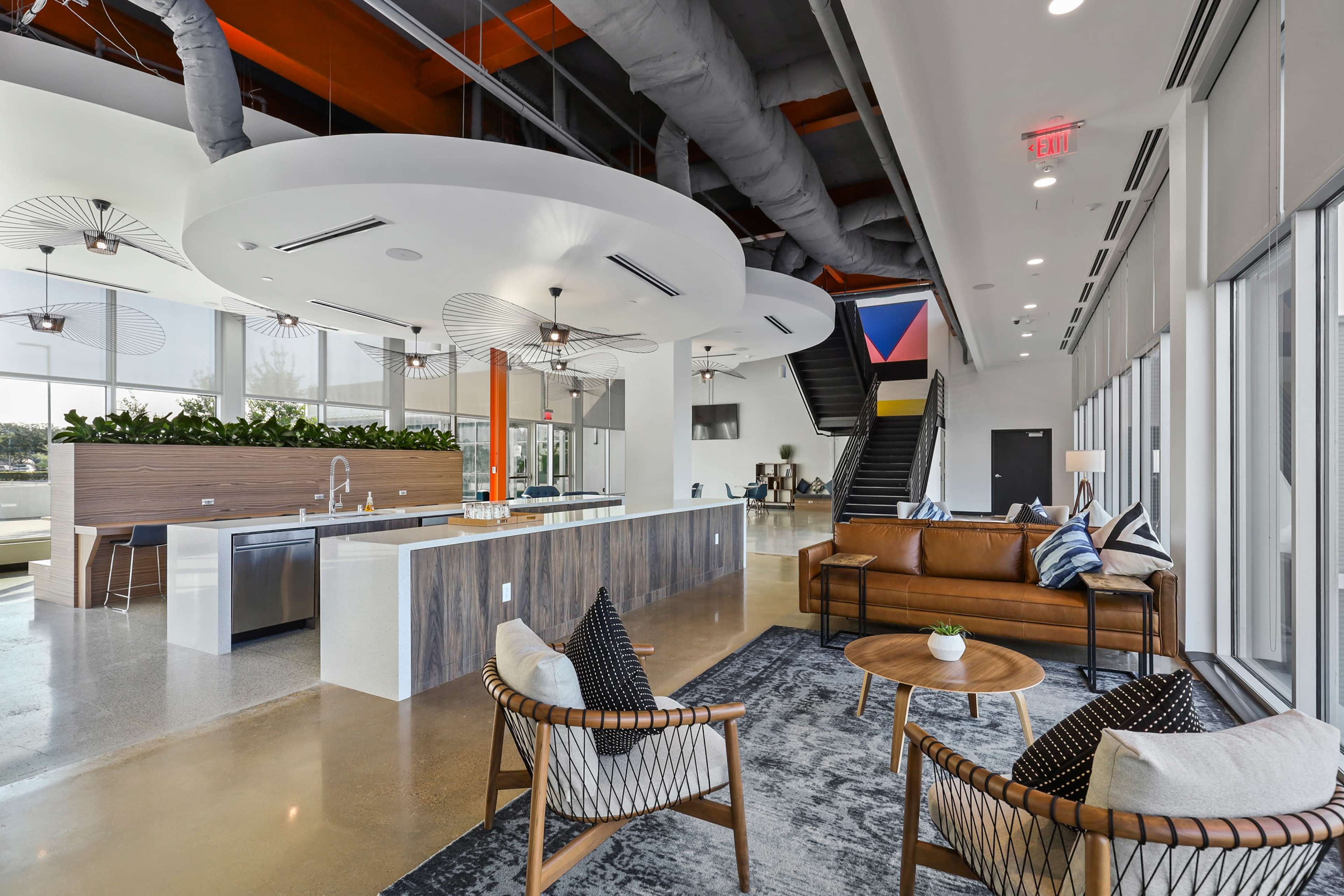 The image shows a modern, open-concept lounge area featuring a kitchen island, seating arrangements, and large windows that provide ample natural light.