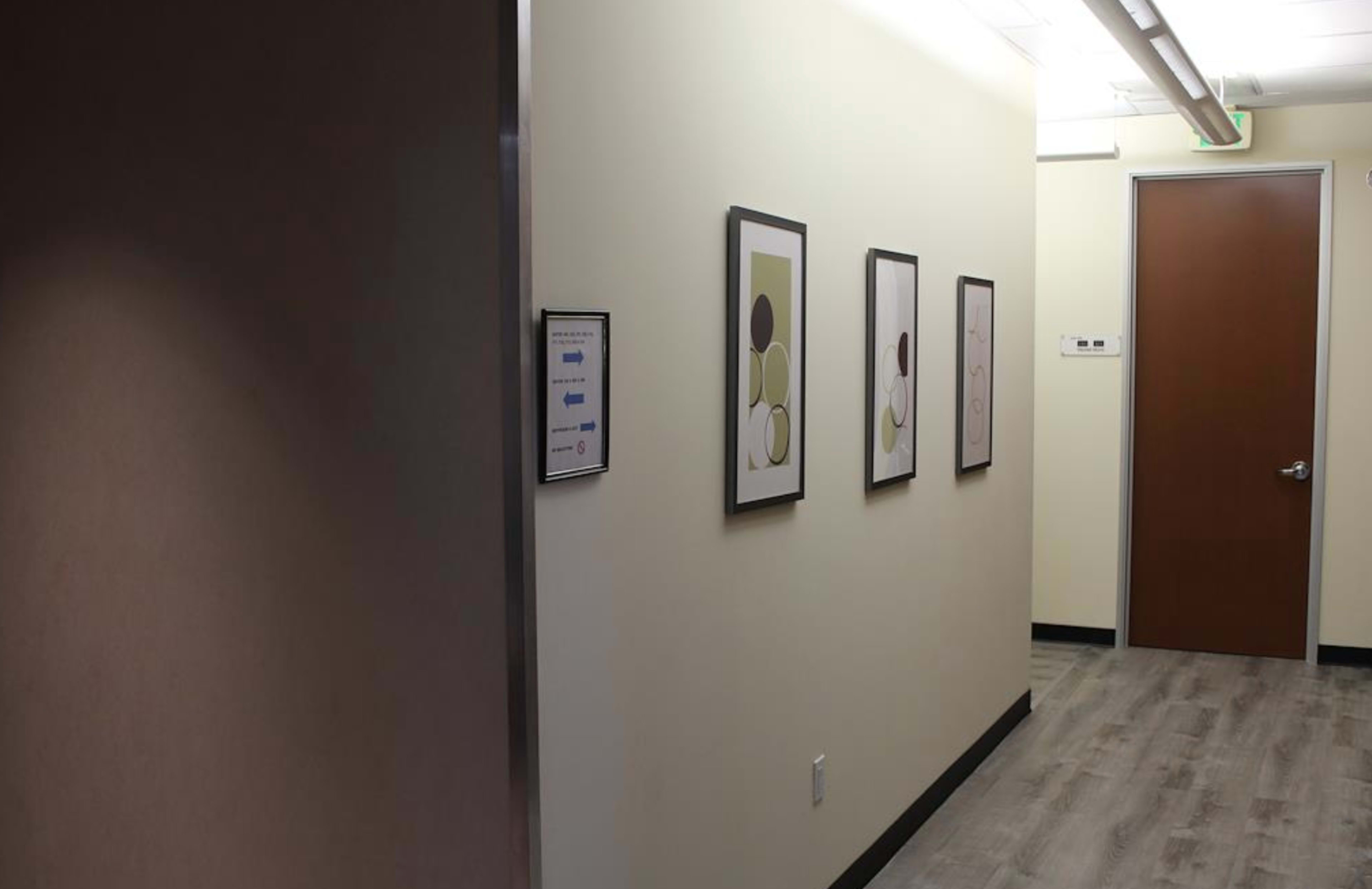 A well-lit hallway with light-colored walls, featuring three framed artworks on one side and a closed door at the far end.