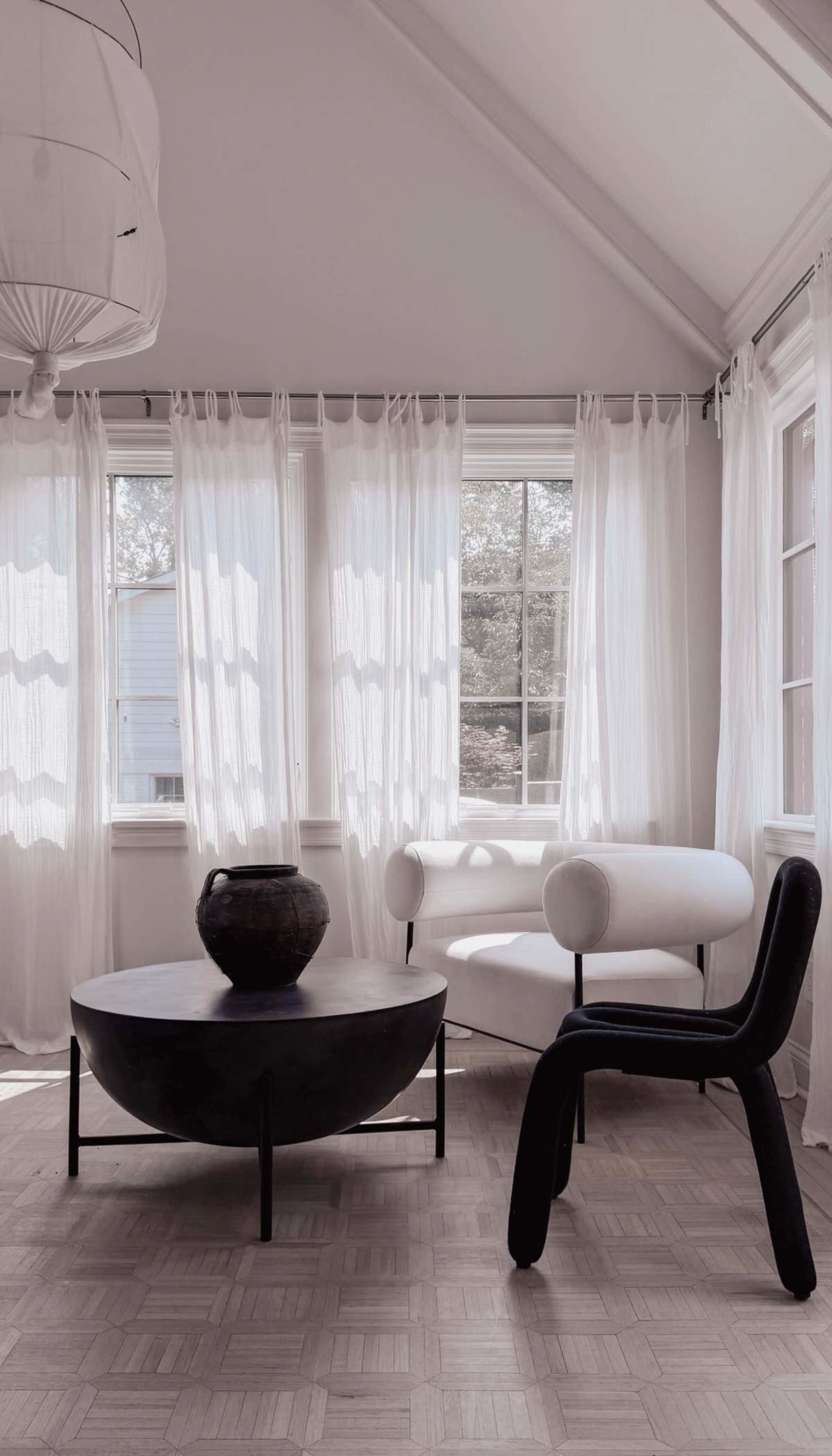 The image depicts a bright, modern living space featuring a round black coffee table, a white armchair, and a black chair, all with large windows dressed in sheer white curtains letting in natural light.