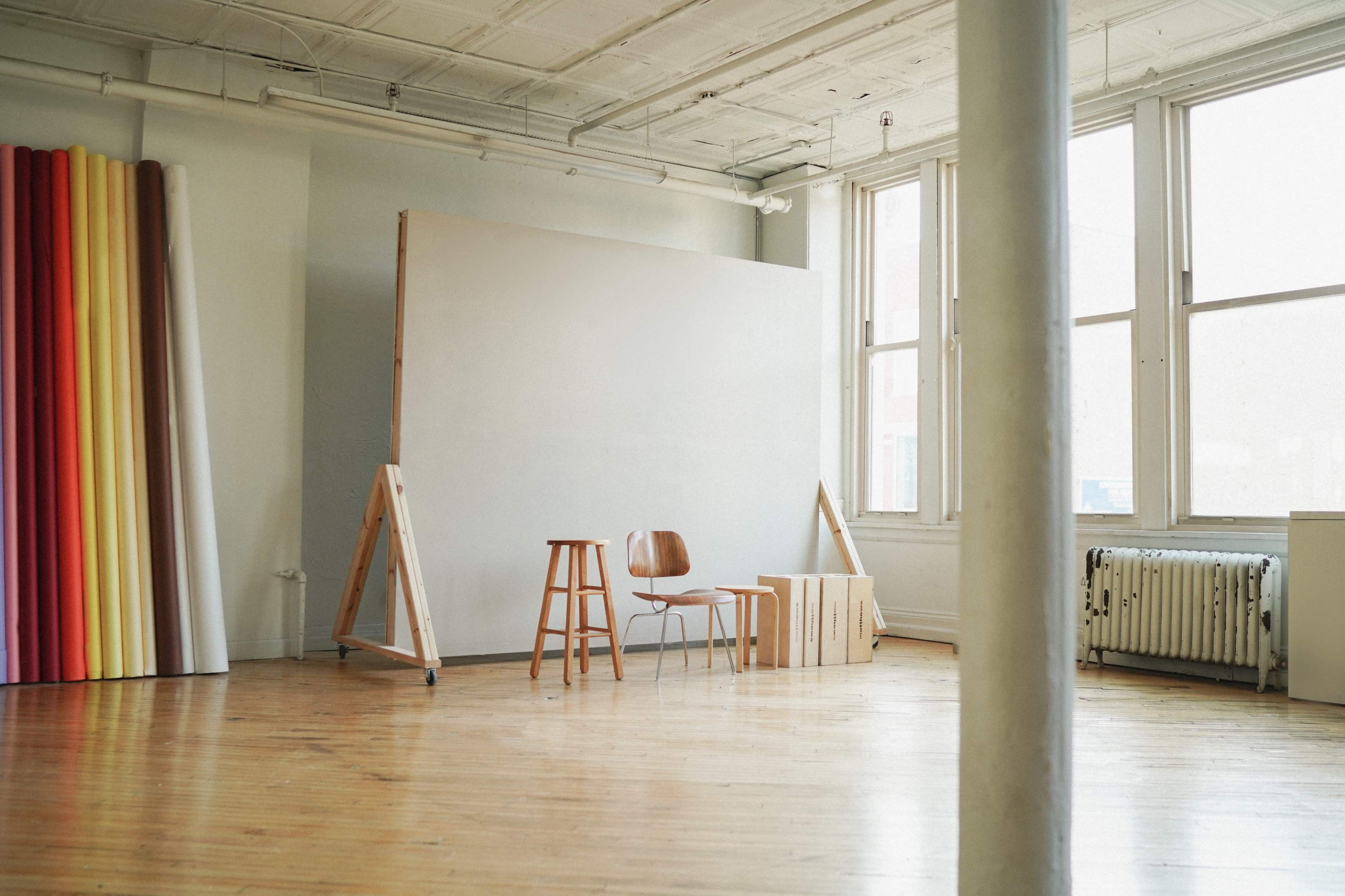 The image shows a minimalist studio space with a blank canvas, two chairs, a wooden crate, and rolls of colored paper against the wall.