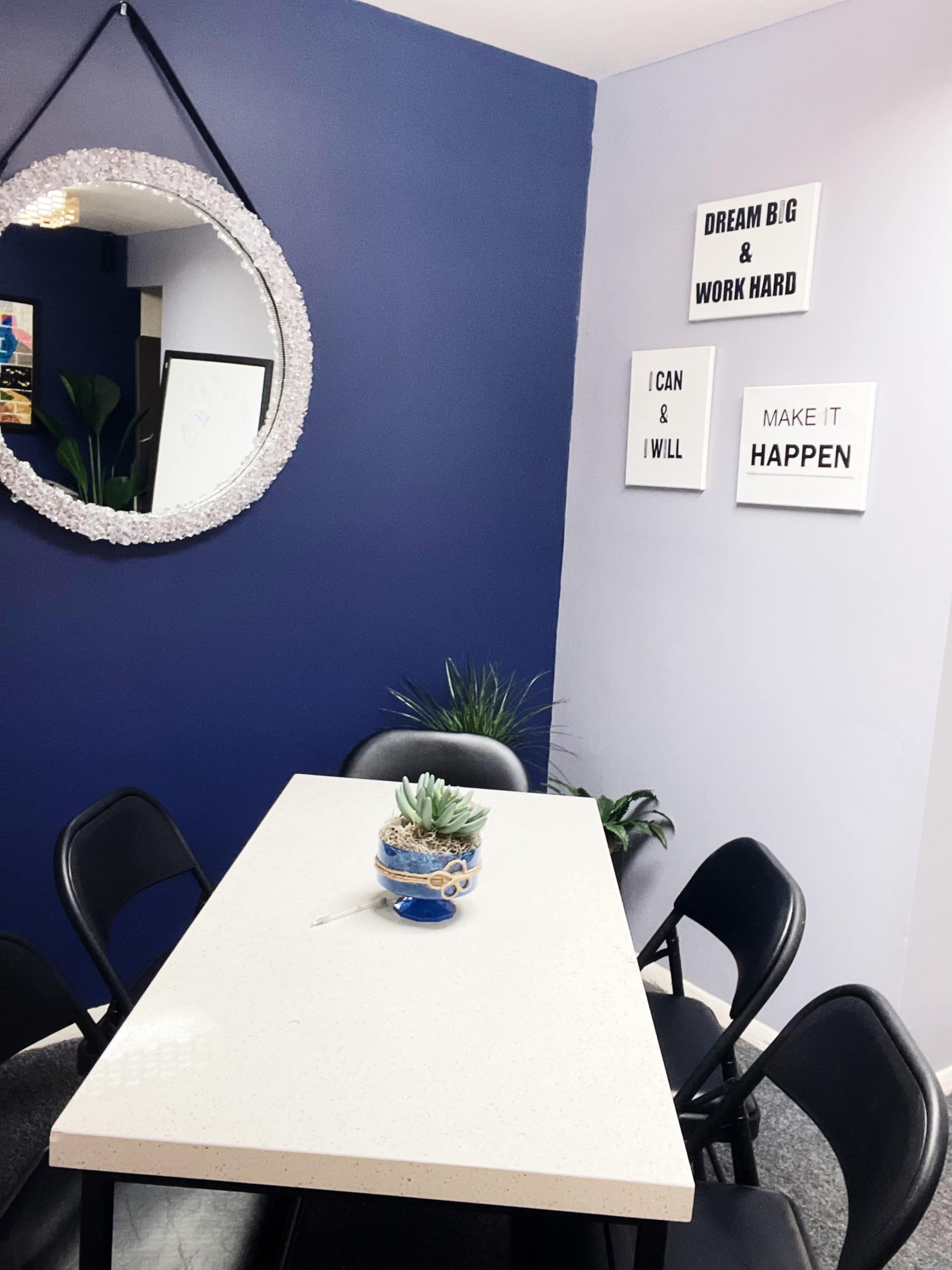 A small dining area features a circular mirror on the wall, a white table surrounded by black chairs, and motivational art on the purple wall.