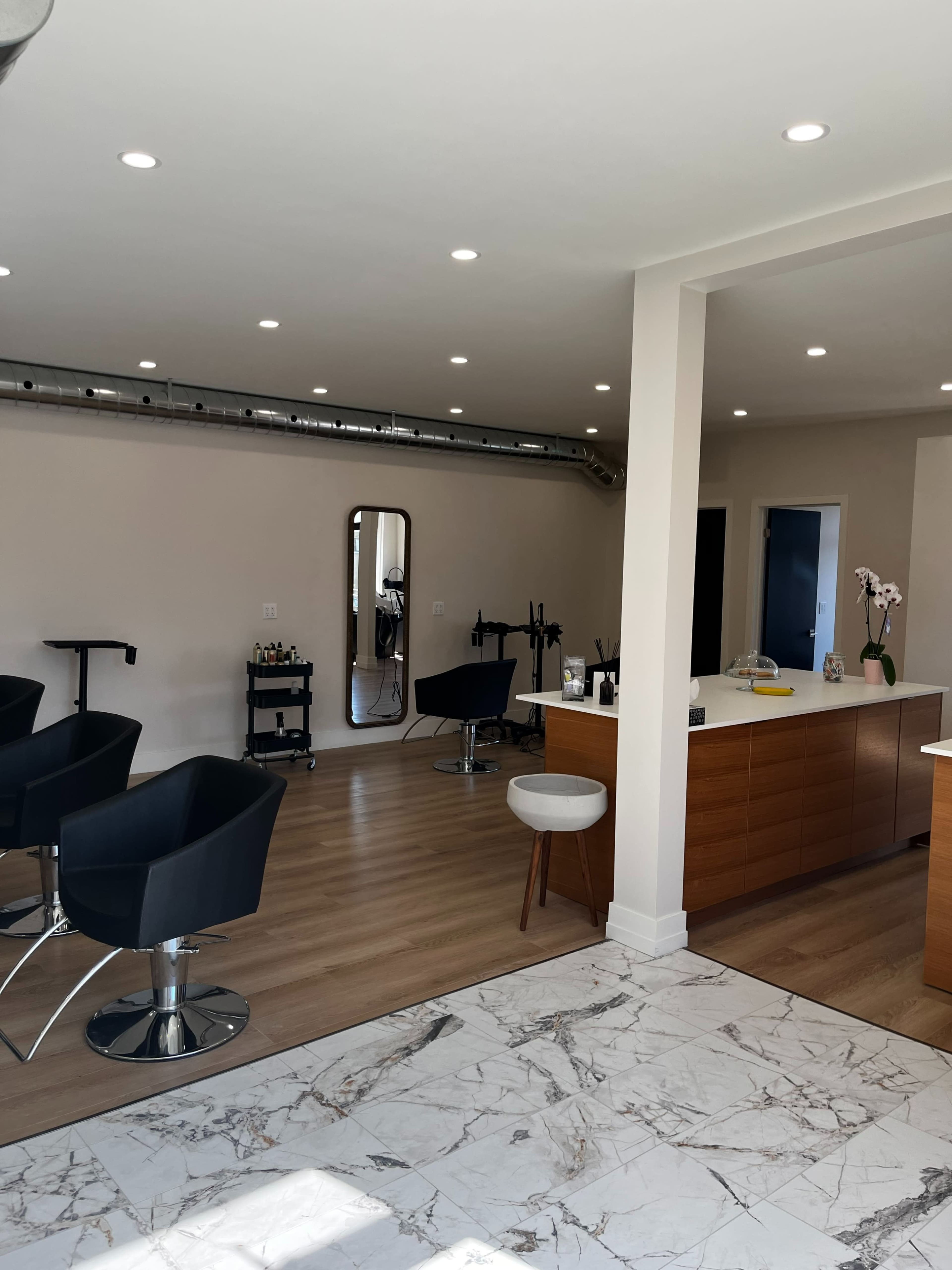 A modern hair salon with black styling chairs, a large mirror, and a reception area featuring a wooden desk.