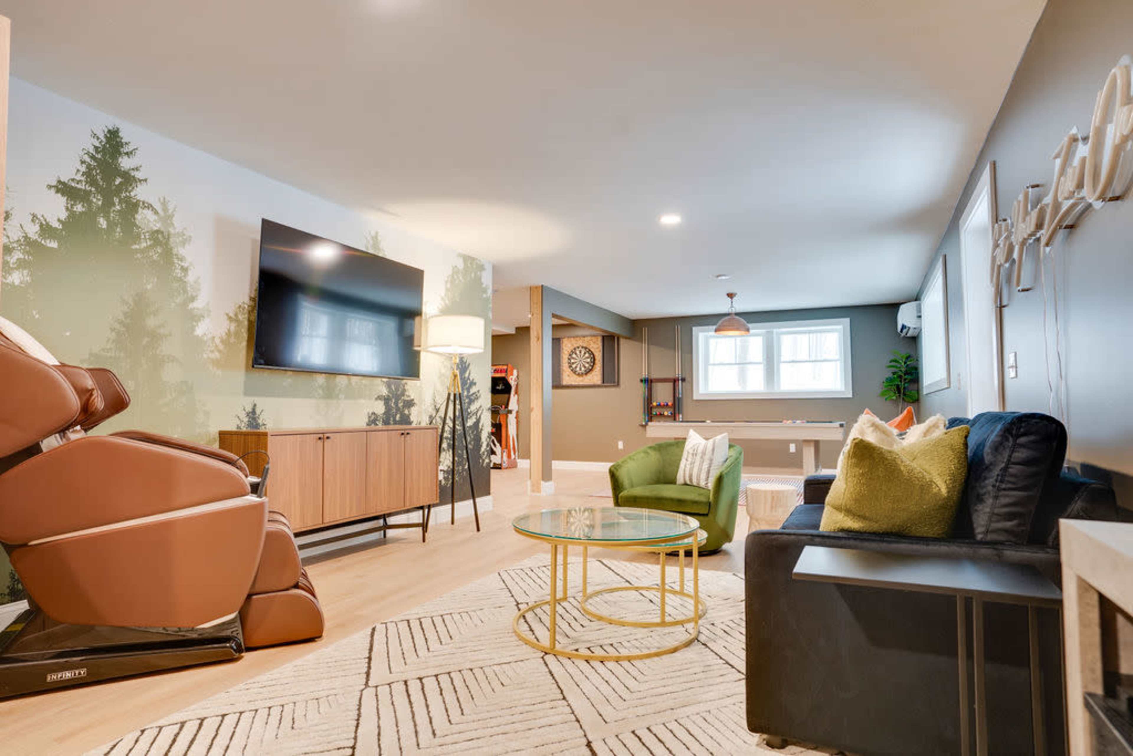 The image shows a modern living room setup in a basement, featuring a green chair, a black sofa, a glass coffee table, a large television, and a wall mural of trees.