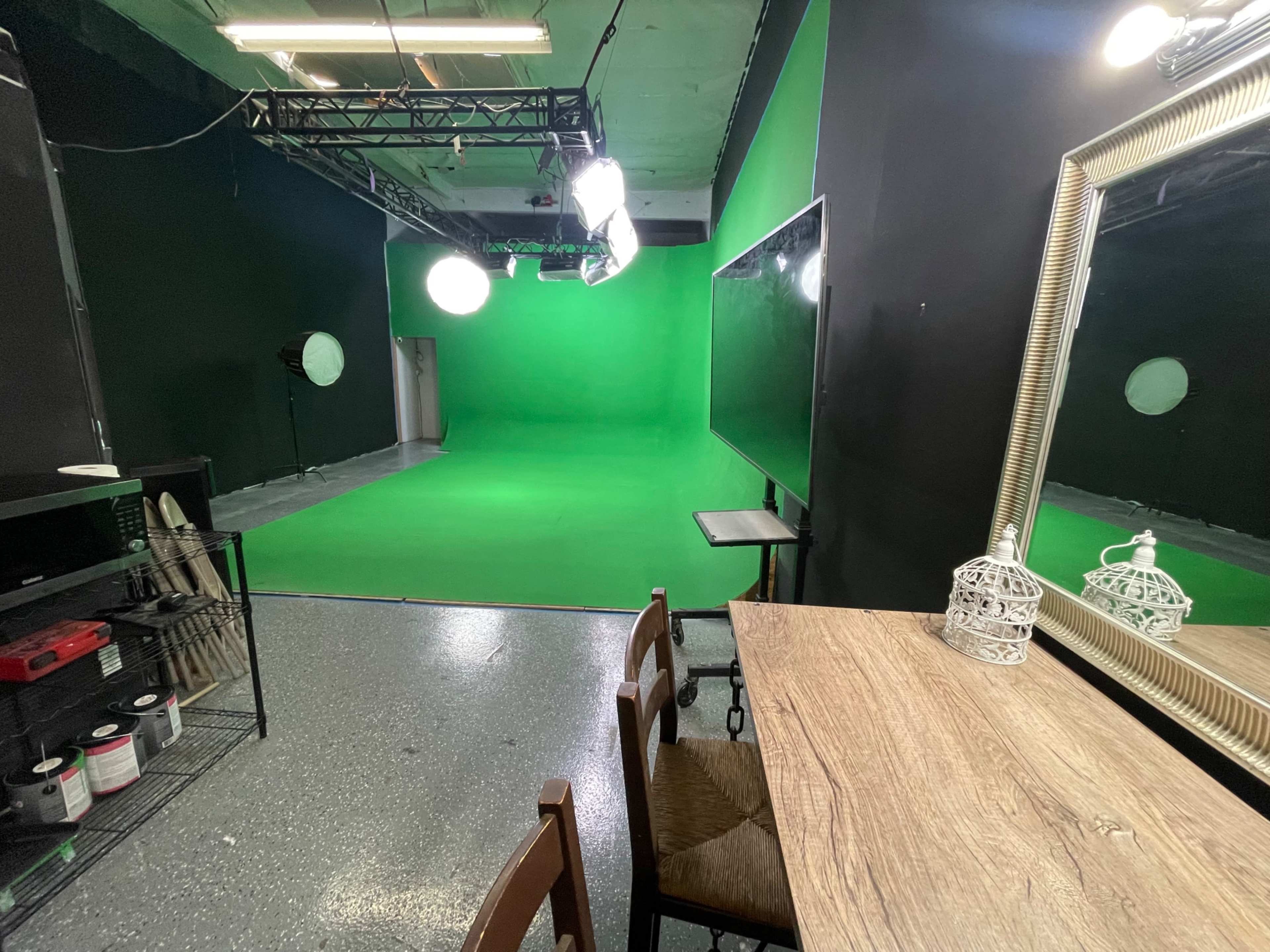 The image shows a green screen studio with lighting equipment set up, a large mirror, and a wooden table with chairs.
