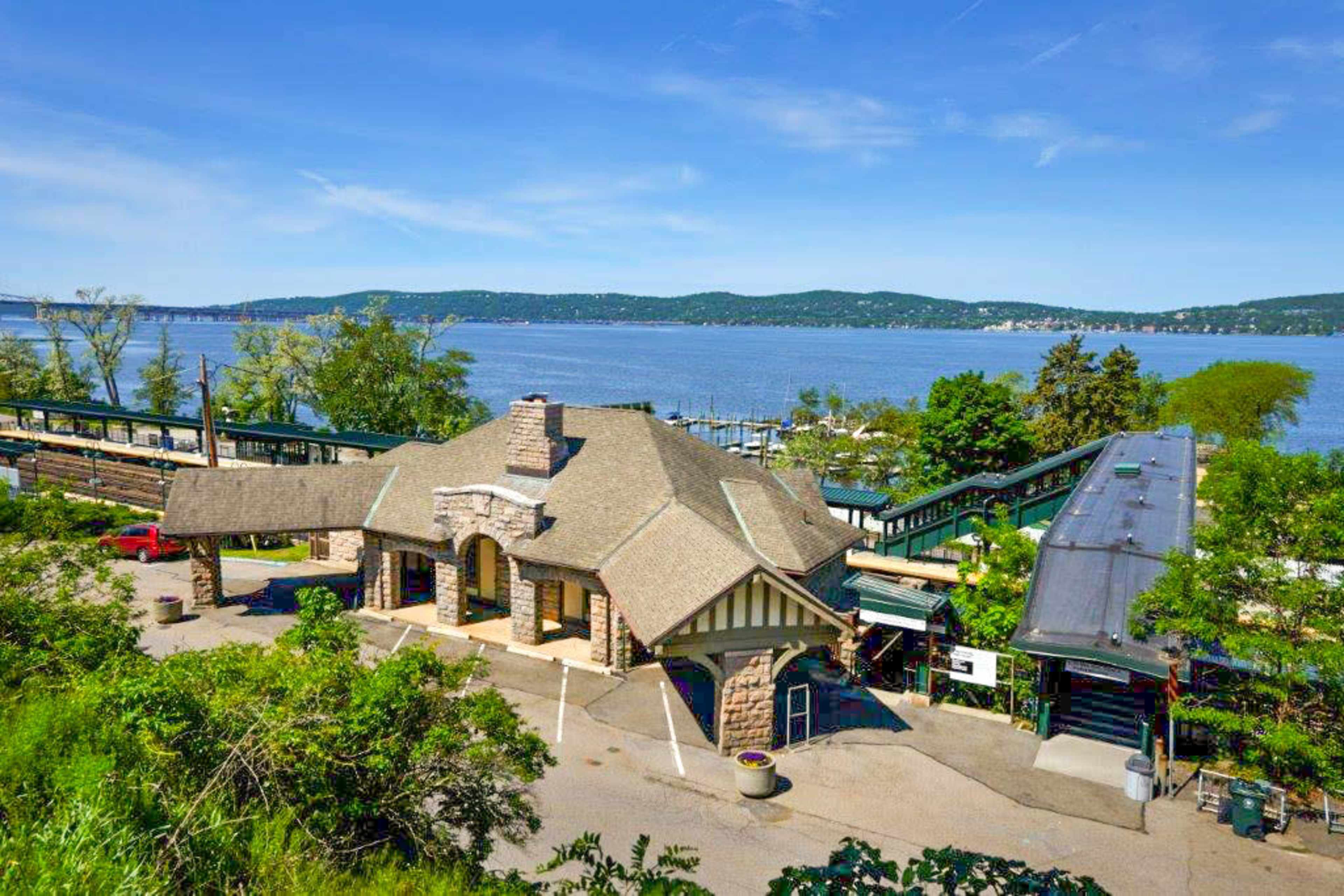 The image shows a waterfront train station with a stone building and a scenic view of the river and hills in the background.