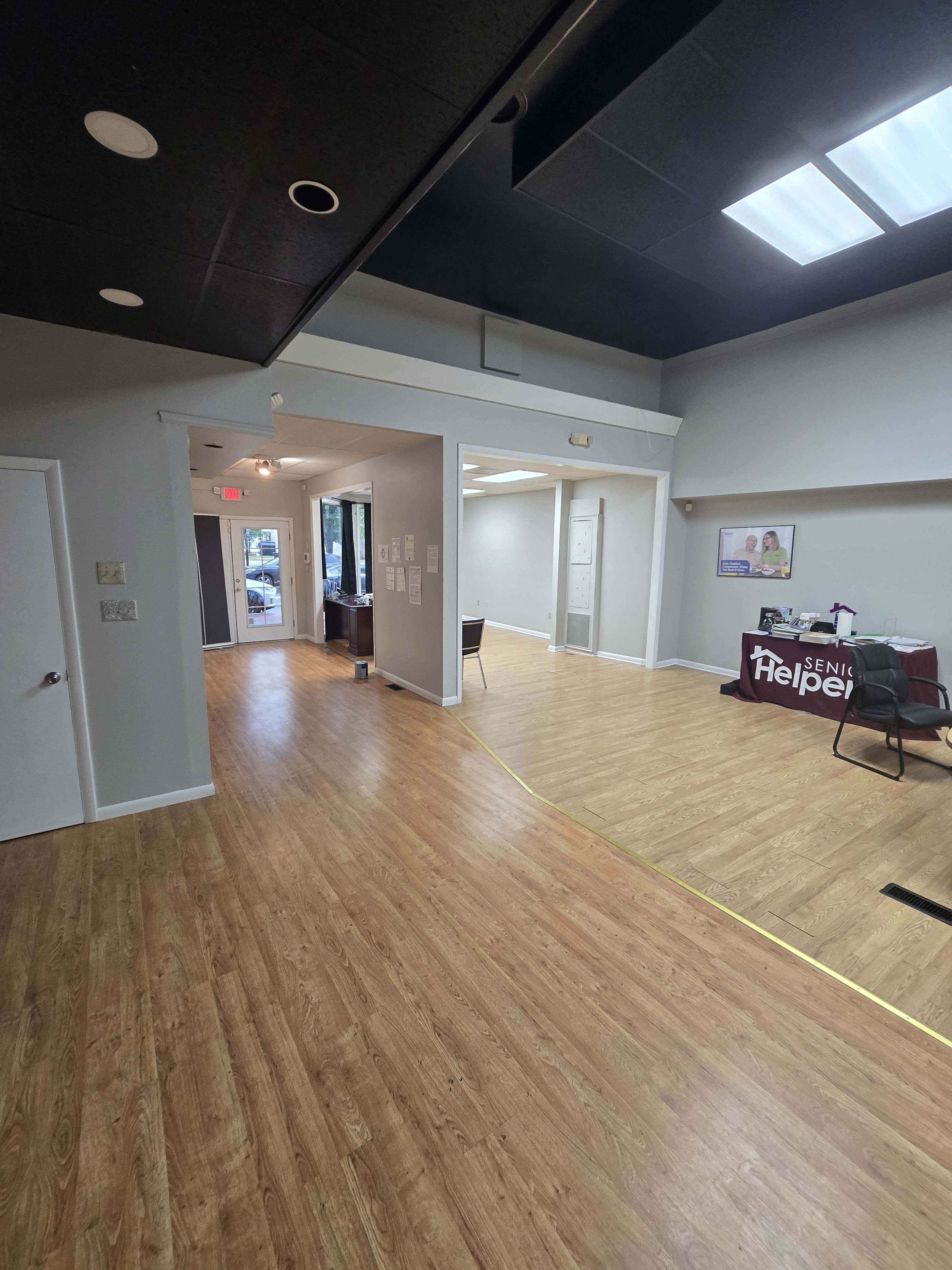 The image shows an interior space with wooden flooring and a divided area, featuring a table with a sign for "Senior Helpers" on one side and an entrance leading to another room.