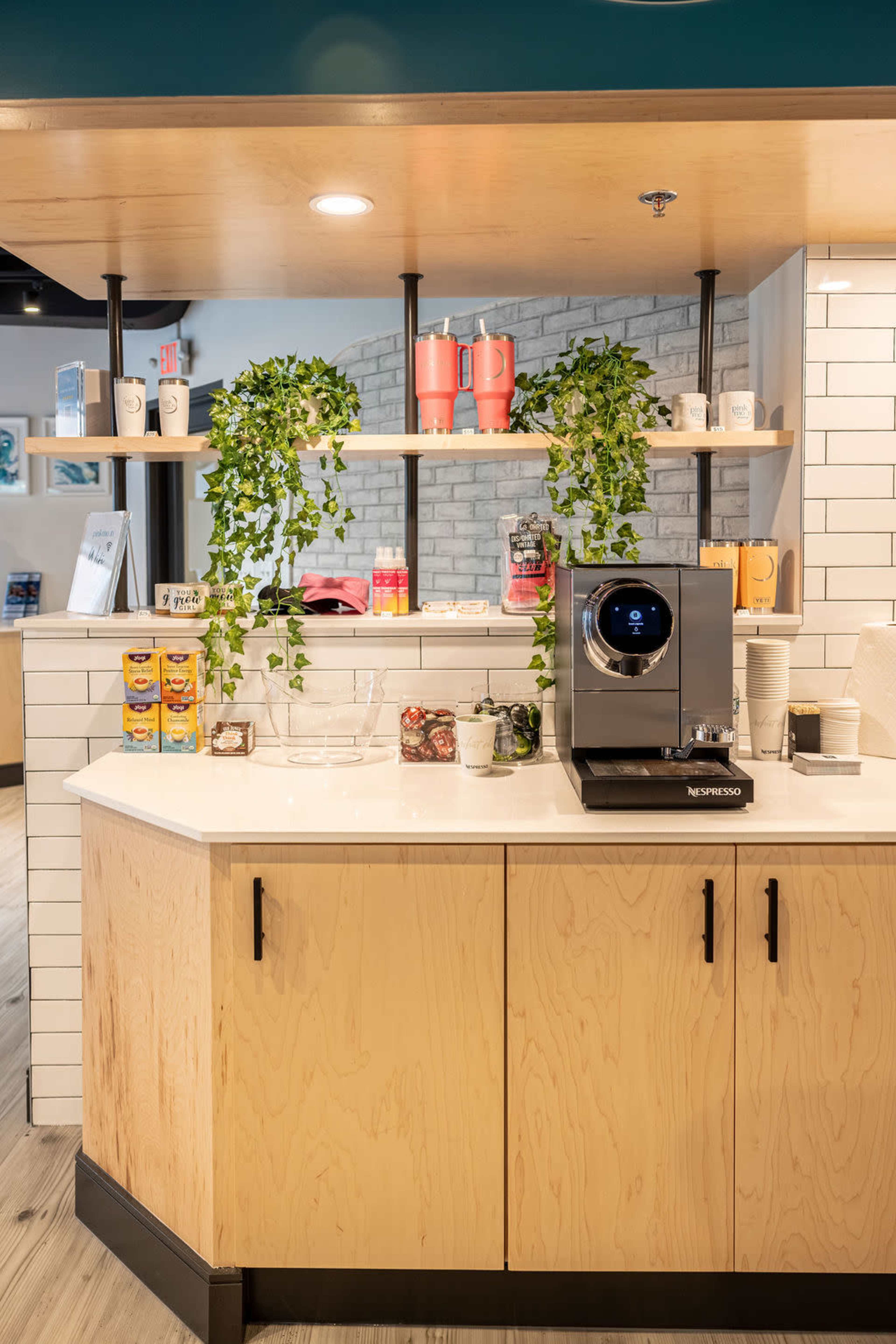 The image shows a modern coffee station with a Nespresso machine, shelves displaying beverages and plants, and a countertop made of light-colored wood.