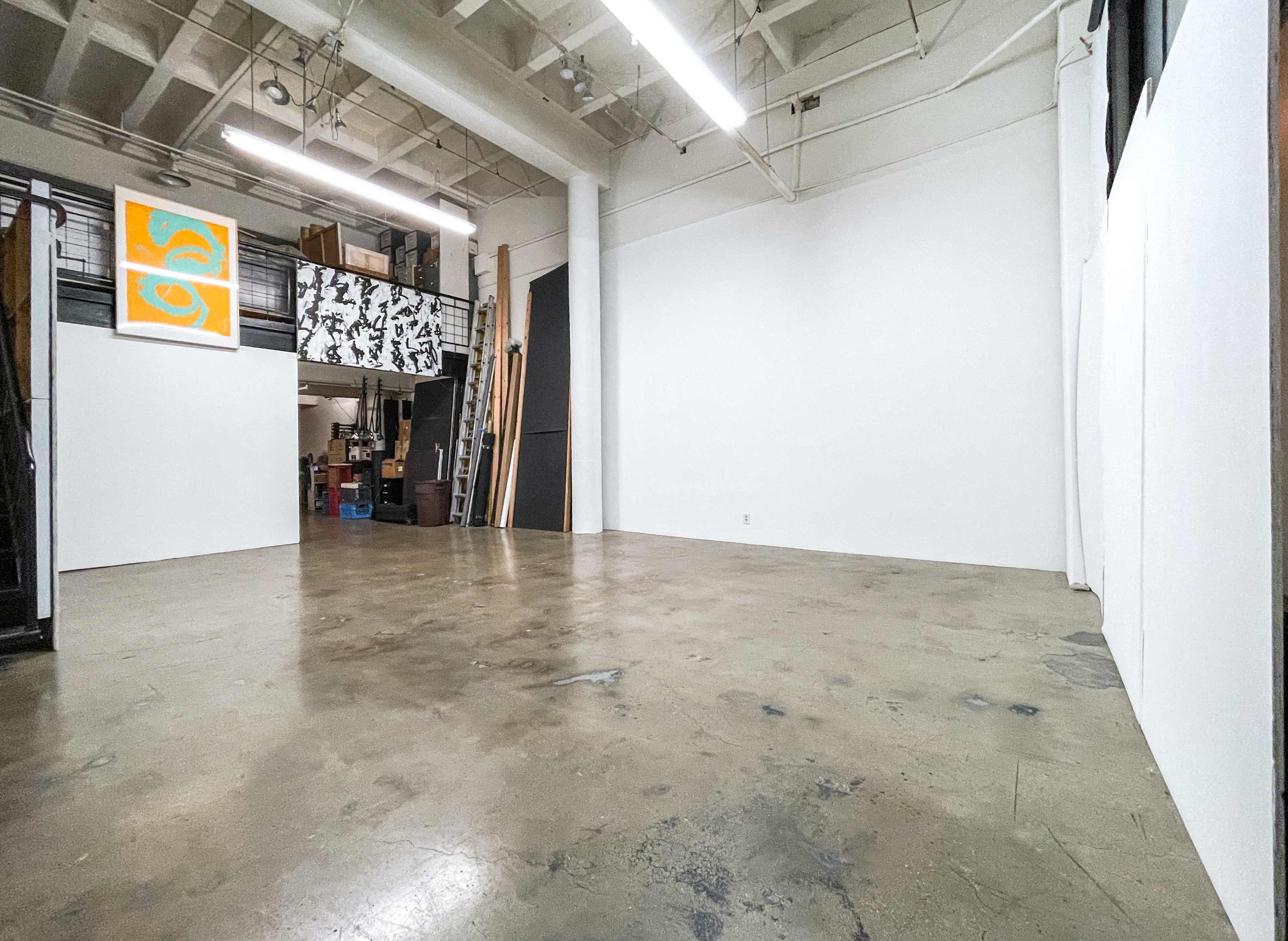 The image shows a spacious, empty room with a polished concrete floor and white walls, featuring art on the upper left and storage materials in the back.