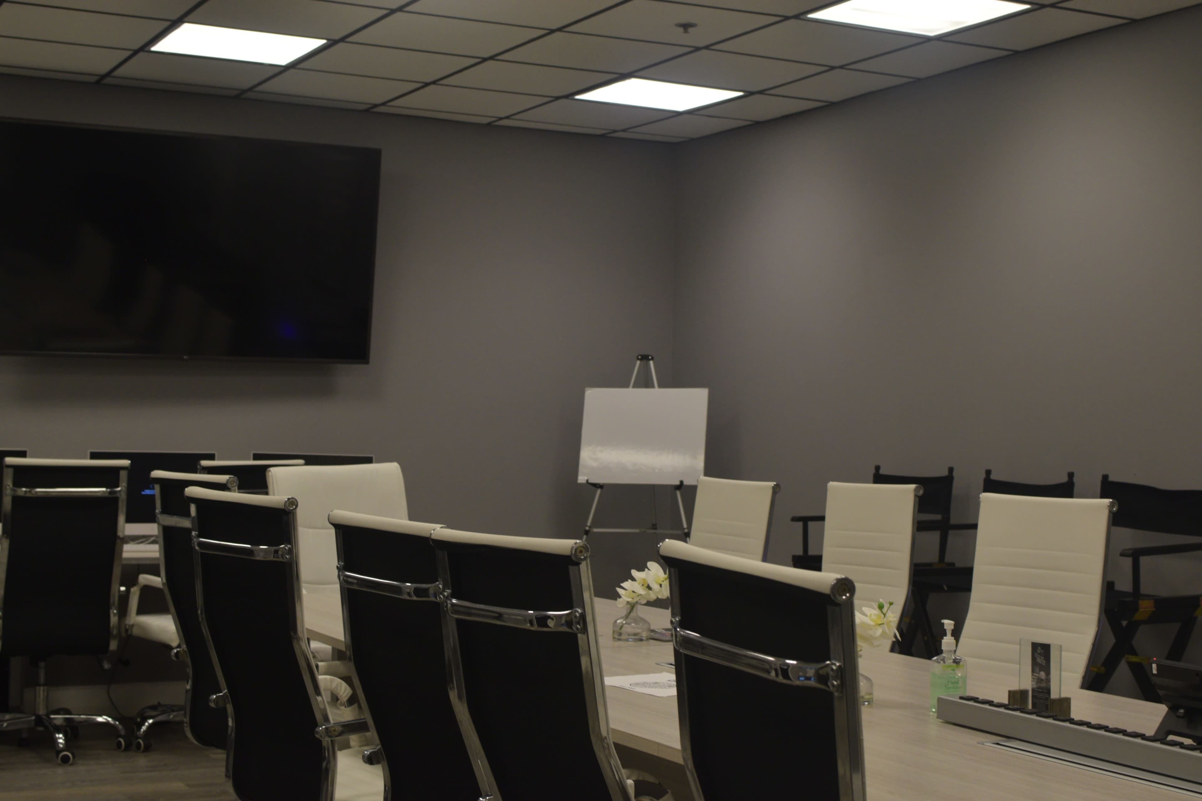 A modern conference room features a long table surrounded by white and black chairs, a large screen on the wall, and an easel with a blank pad.