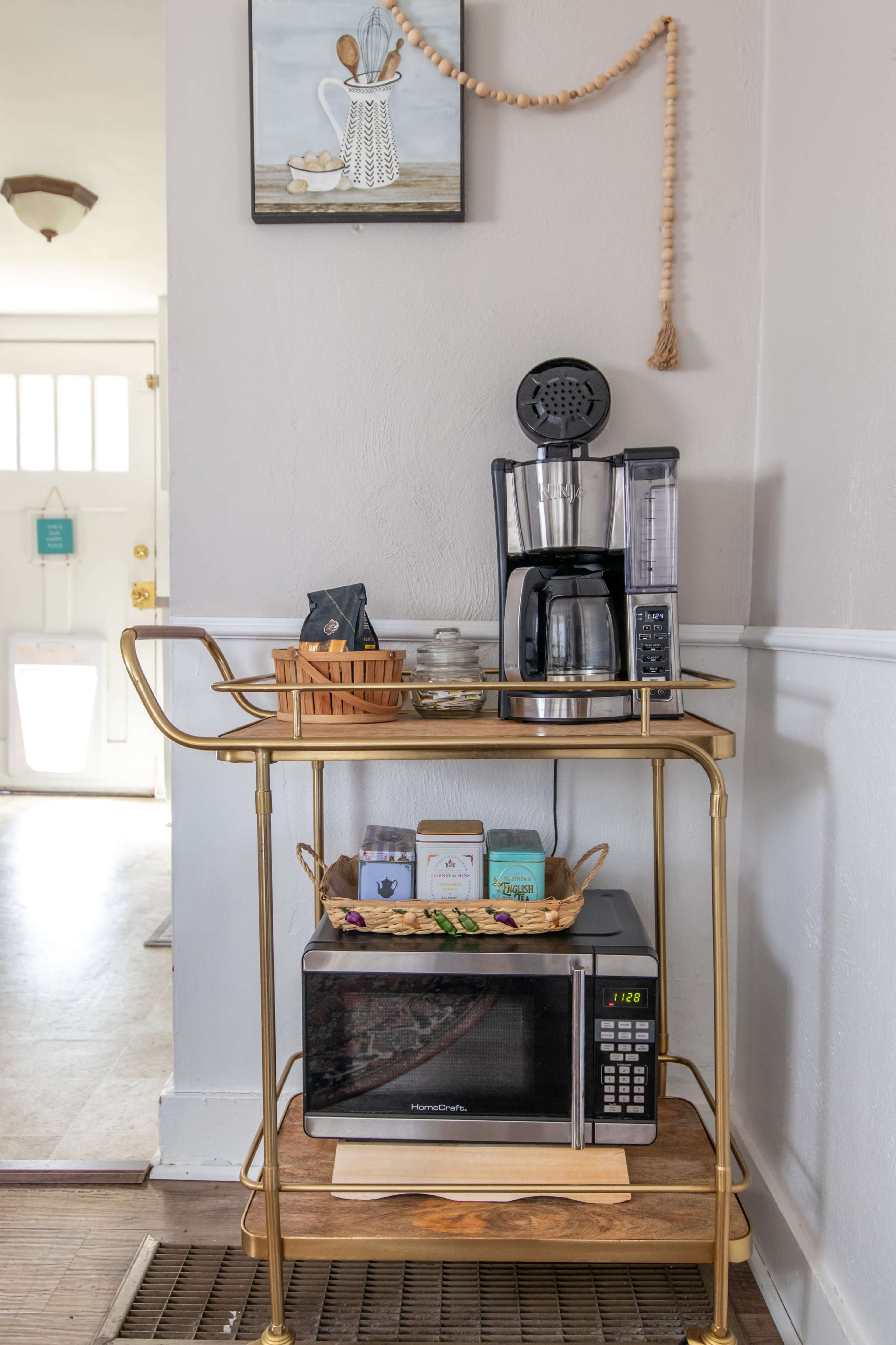 A brass serving cart holds a coffee maker, a microwave, and various items on a wooden shelf, situated in a brightly lit entryway.