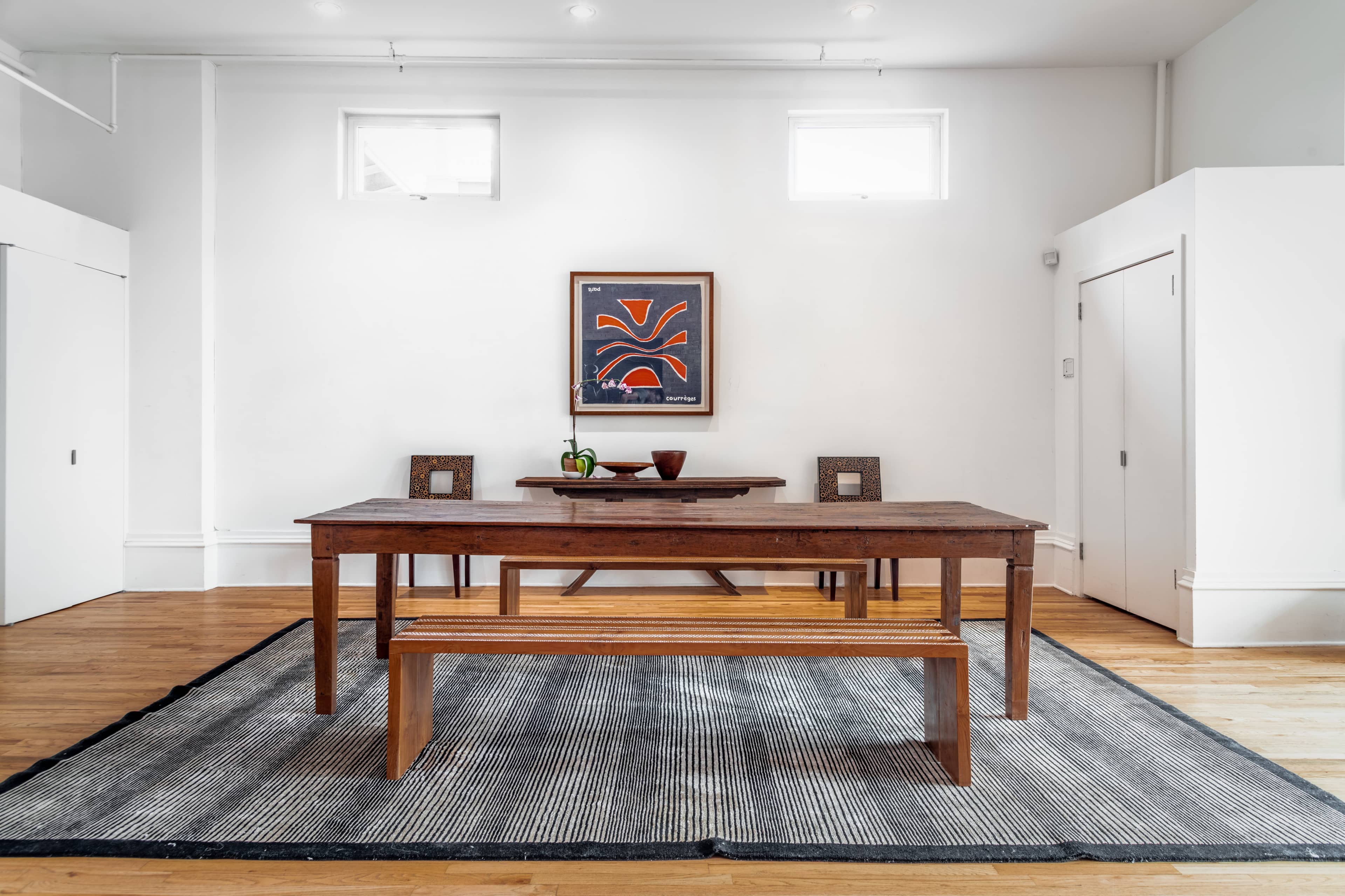 A spacious dining area features a wooden table and bench on a striped rug, with a colorful artwork hanging on the white wall.