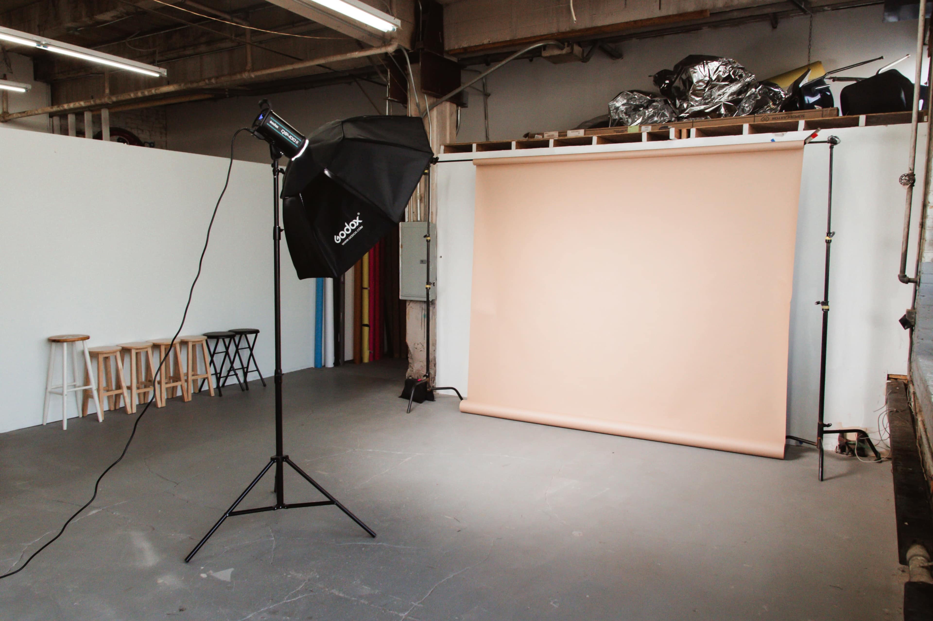 A photography studio with a beige backdrop, a light stand with a softbox, and stools arranged against a wall.