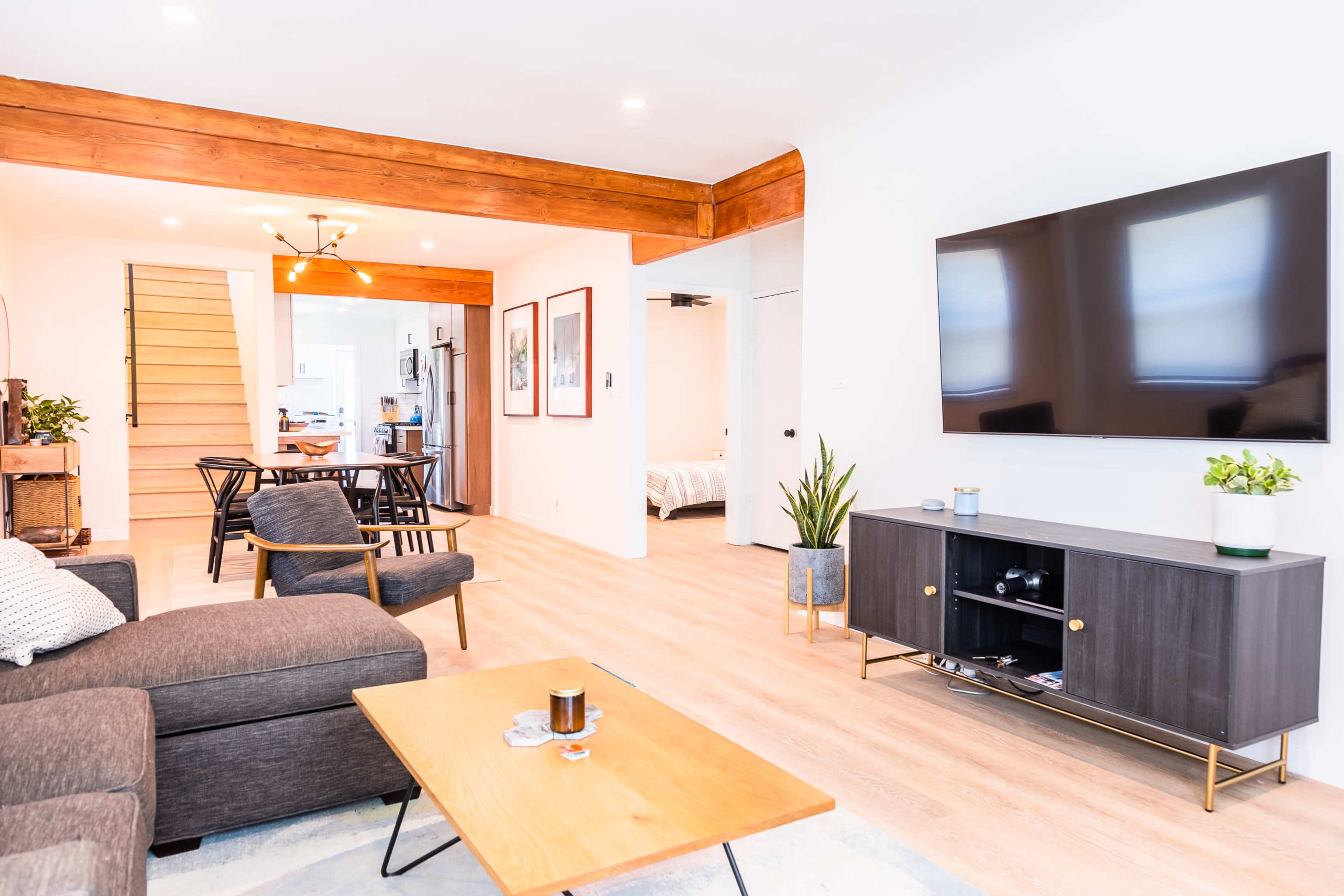 The image shows a modern living room with a sofa, coffee table, and a wall-mounted television, featuring wooden beams and a staircase in the background.