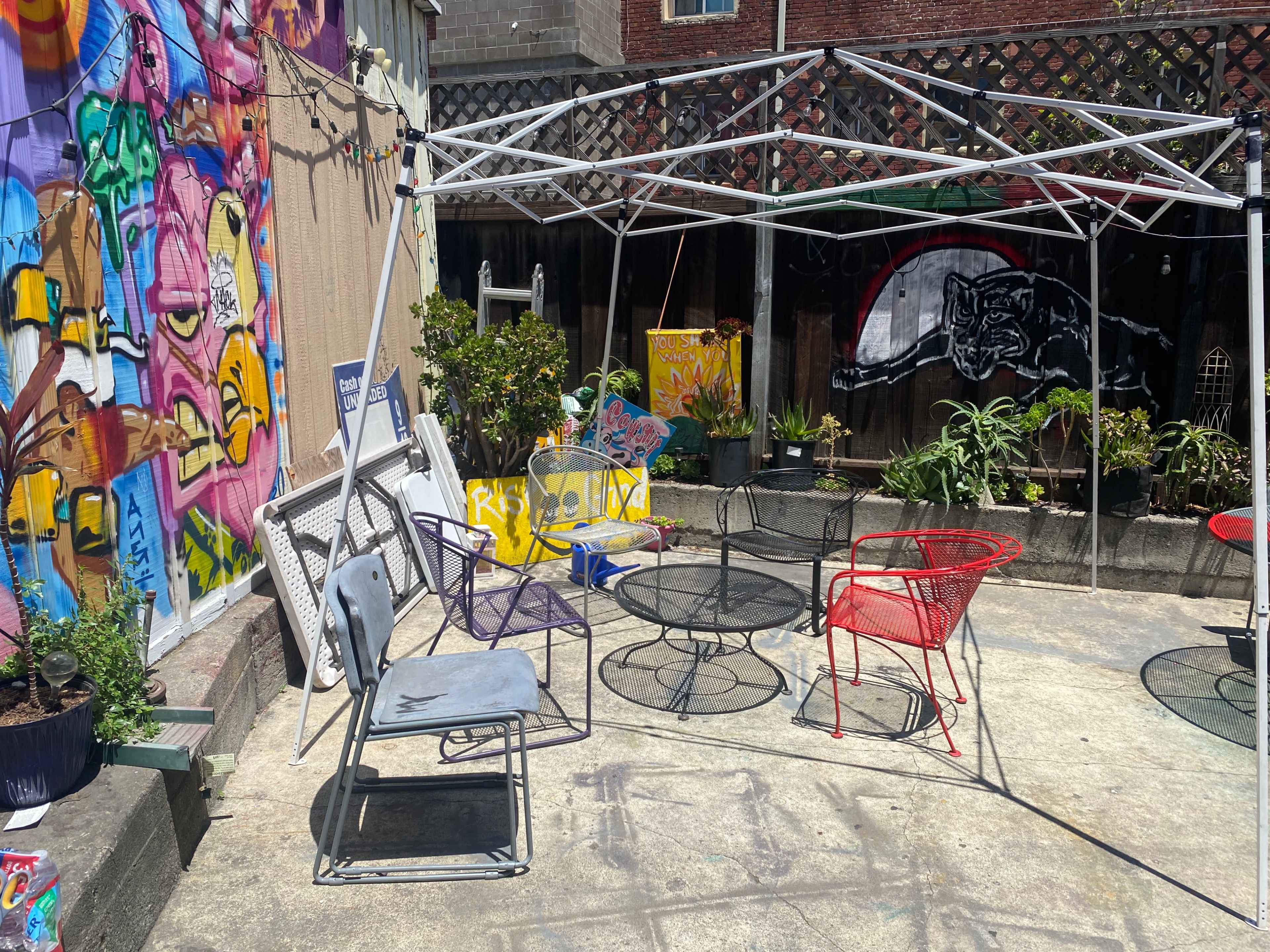 A small outdoor seating area features various chairs and tables under a partially collapsed canopy, surrounded by colorful murals and plants.