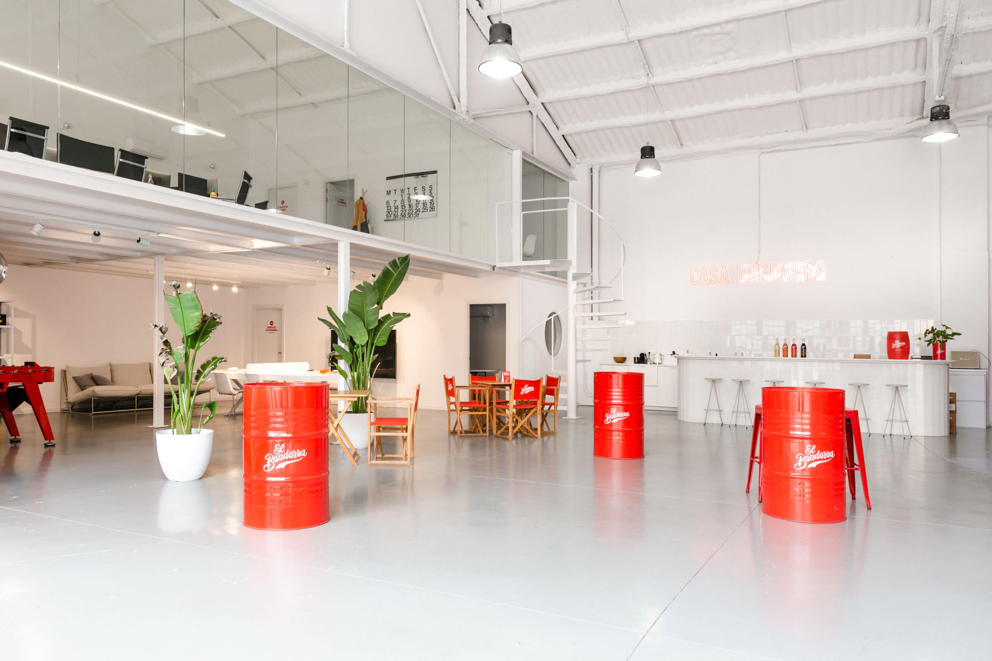 A modern, open-concept workspace featuring red industrial barrels as decor, surrounded by plants and furnished with a combination of seating areas and a kitchen.