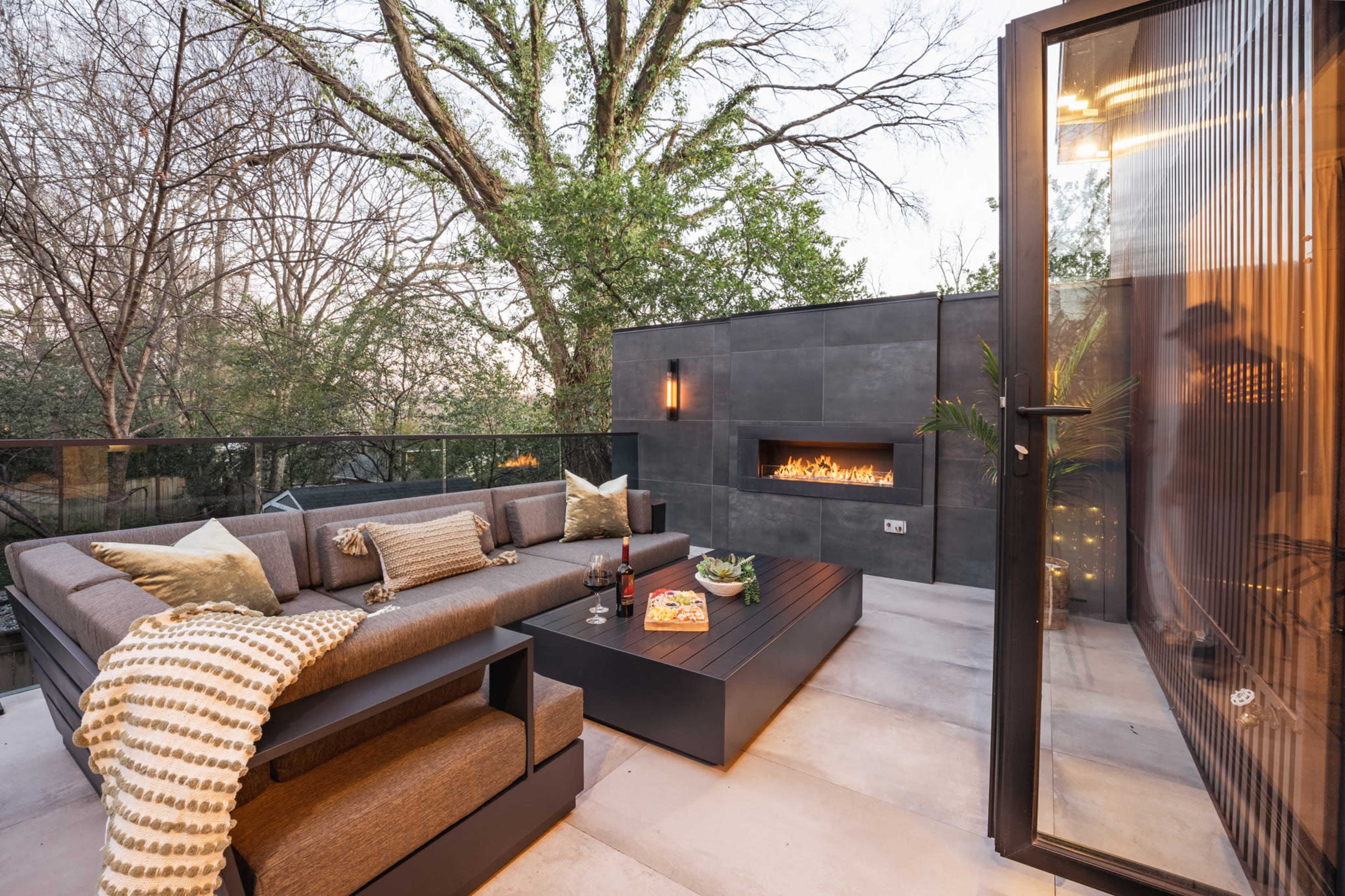 A modern outdoor patio features a comfortable seating area with a fire pit and a view of surrounding trees.