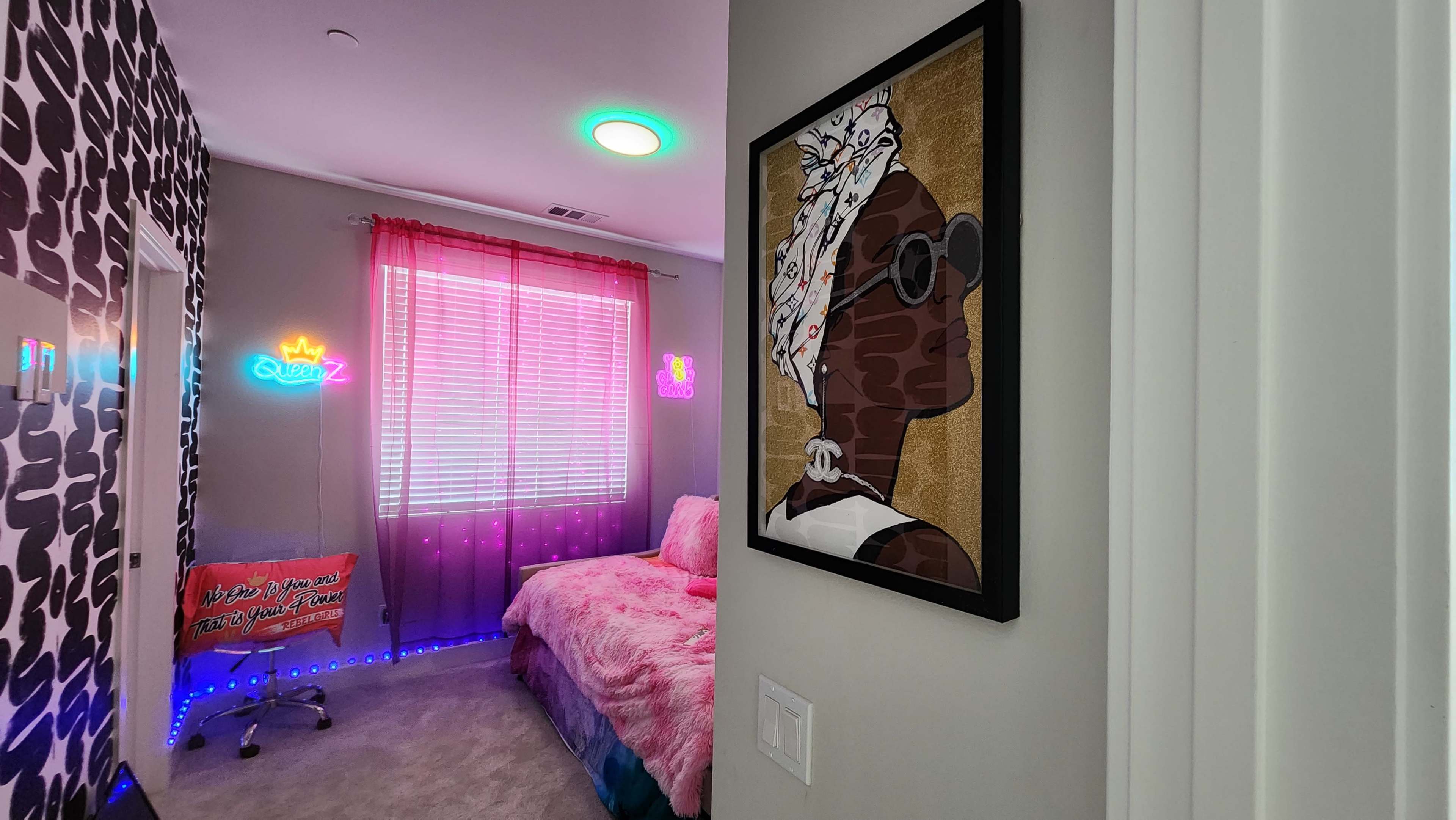 A brightly lit, modern bedroom with a colorful bedspread, decorative wall art, and neon signs.