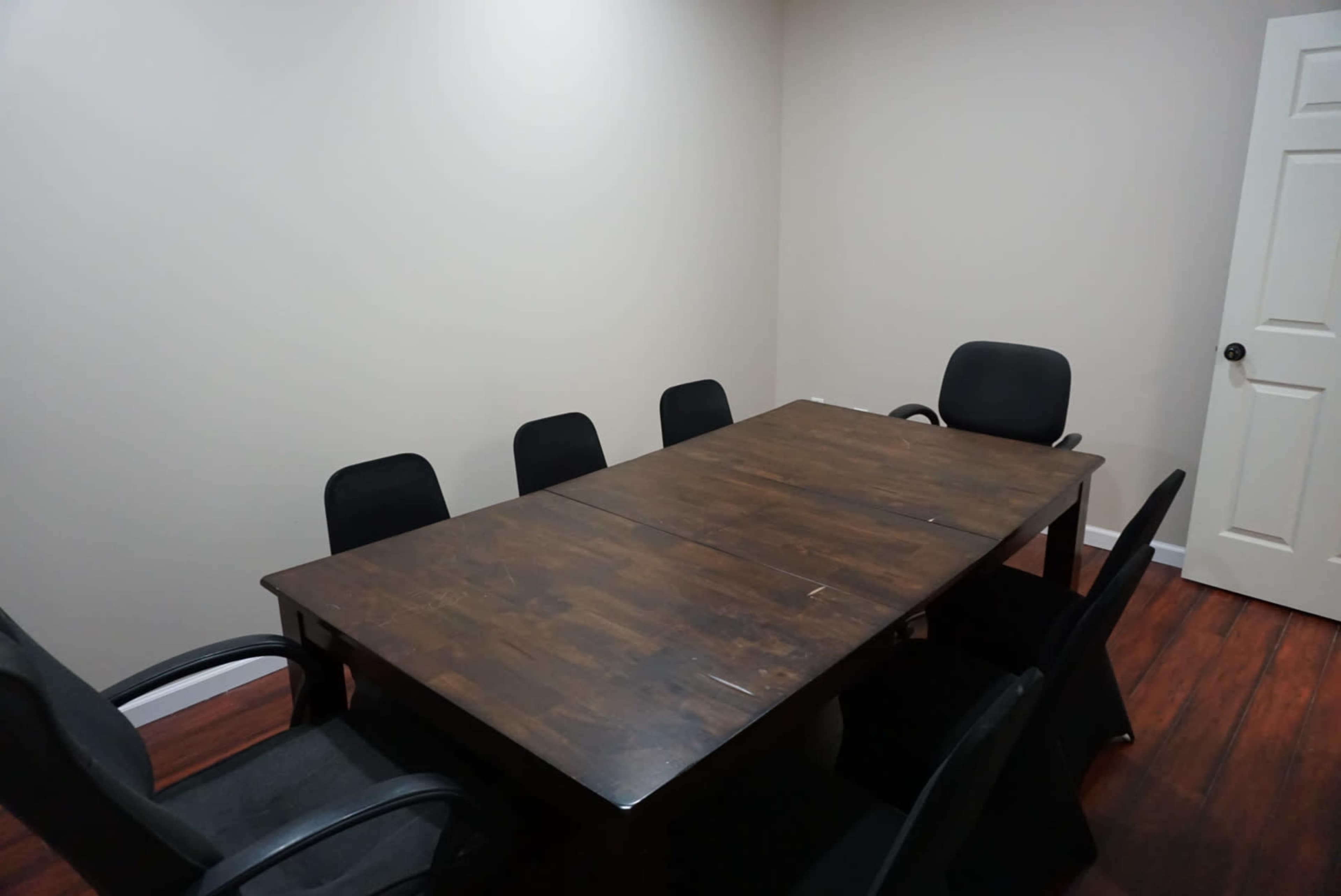 A rectangular wooden table is surrounded by black chairs in a small, empty meeting room with light-colored walls and a single door.