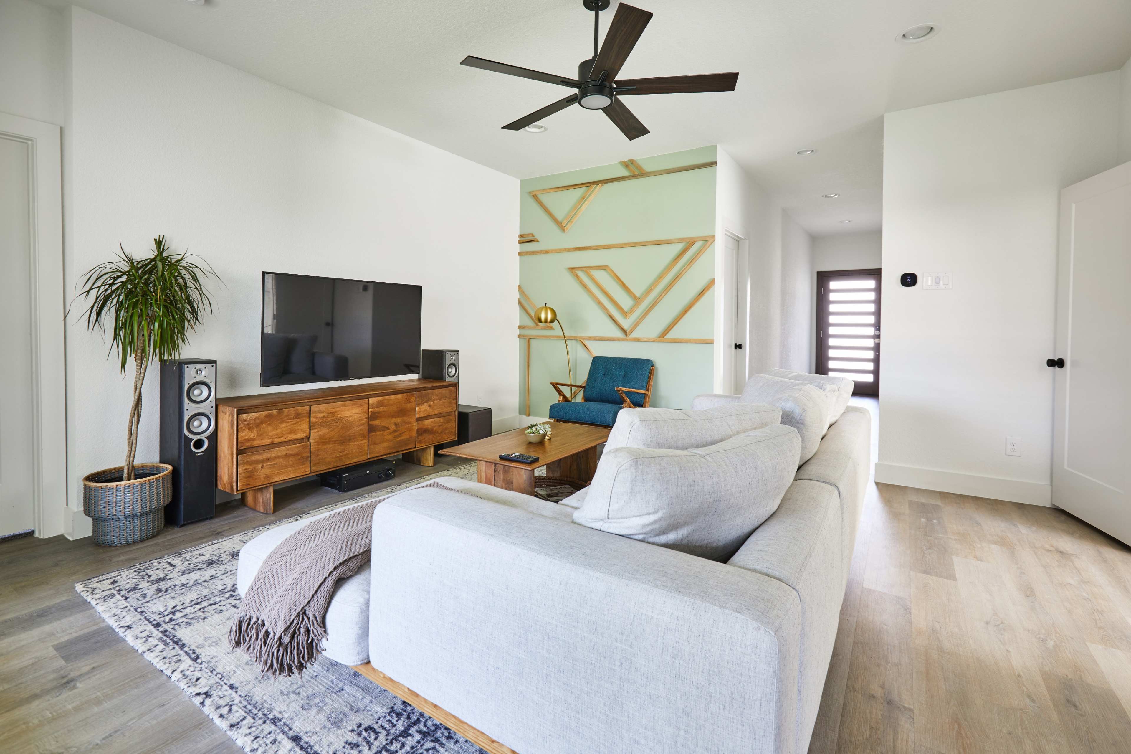 A modern living room features a gray sofa, wooden TV stand, large television, and a mid-century chair against a light green accent wall with geometric decor.