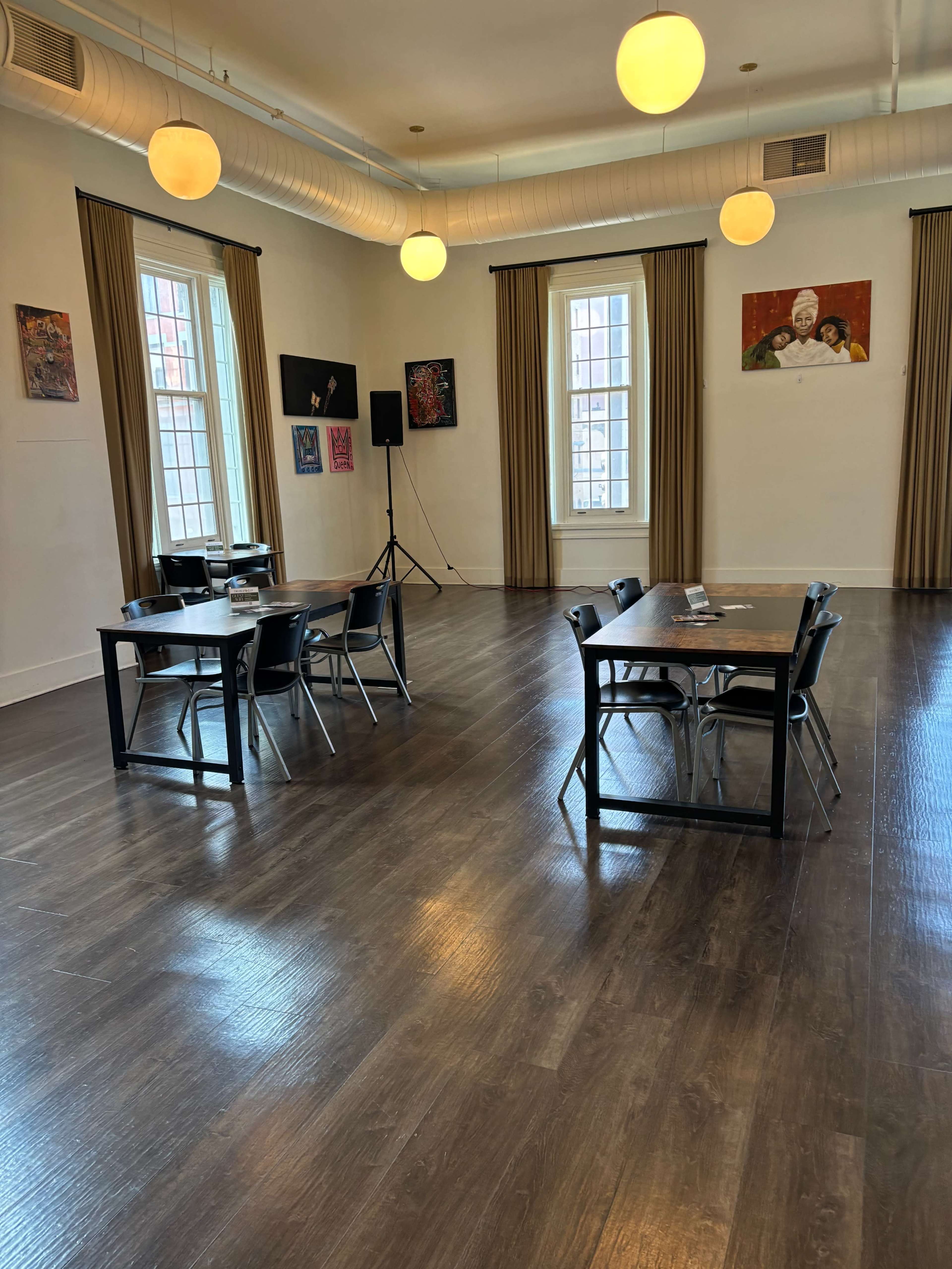 A spacious room with wooden floors, two tables set up with chairs, large windows, and art displayed on the walls.