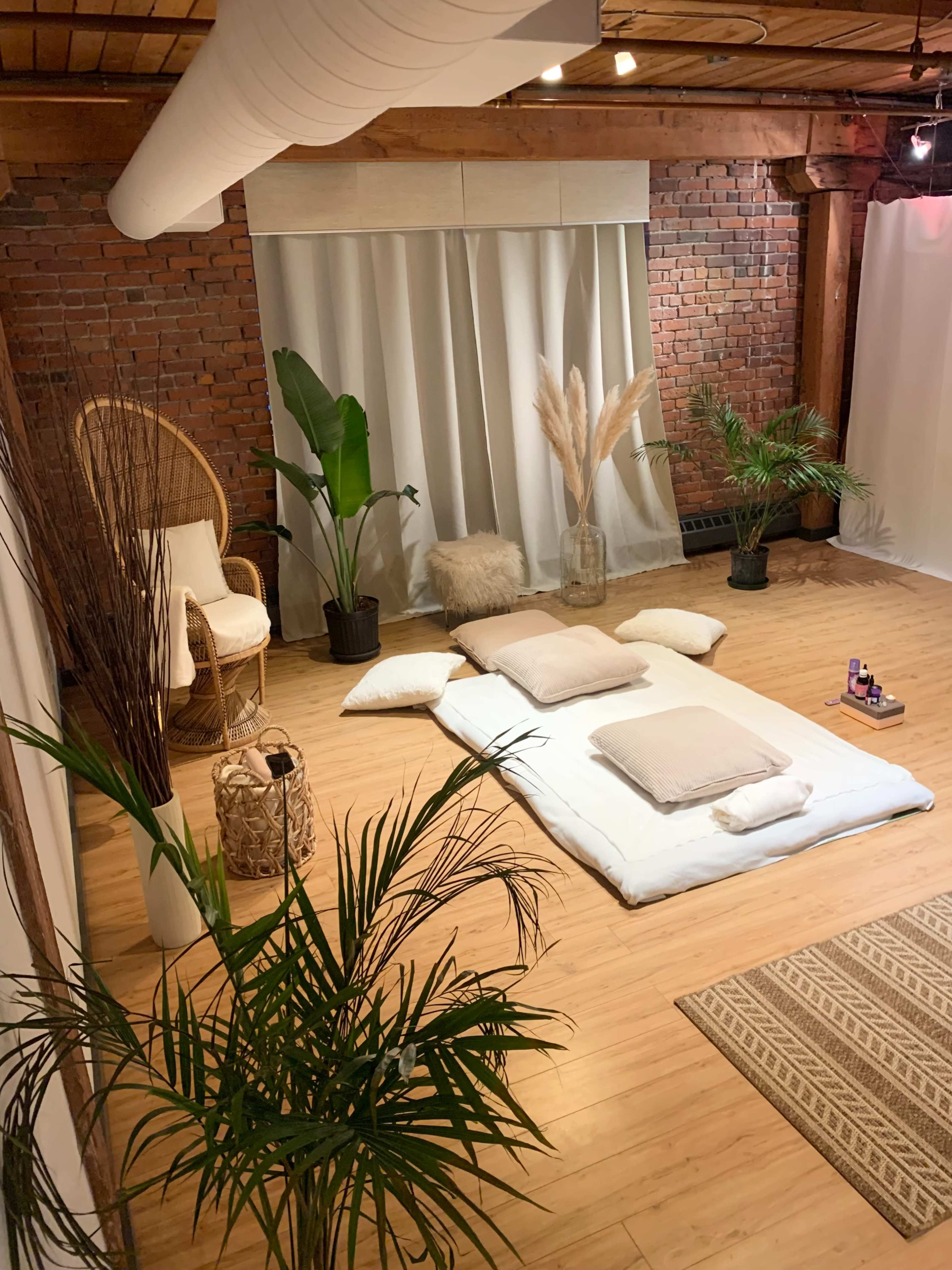 The scene features a cozy minimalist space with a large white mat on the wooden floor, surrounded by plants and soft cushions, and a backdrop of draped fabric and exposed brick.