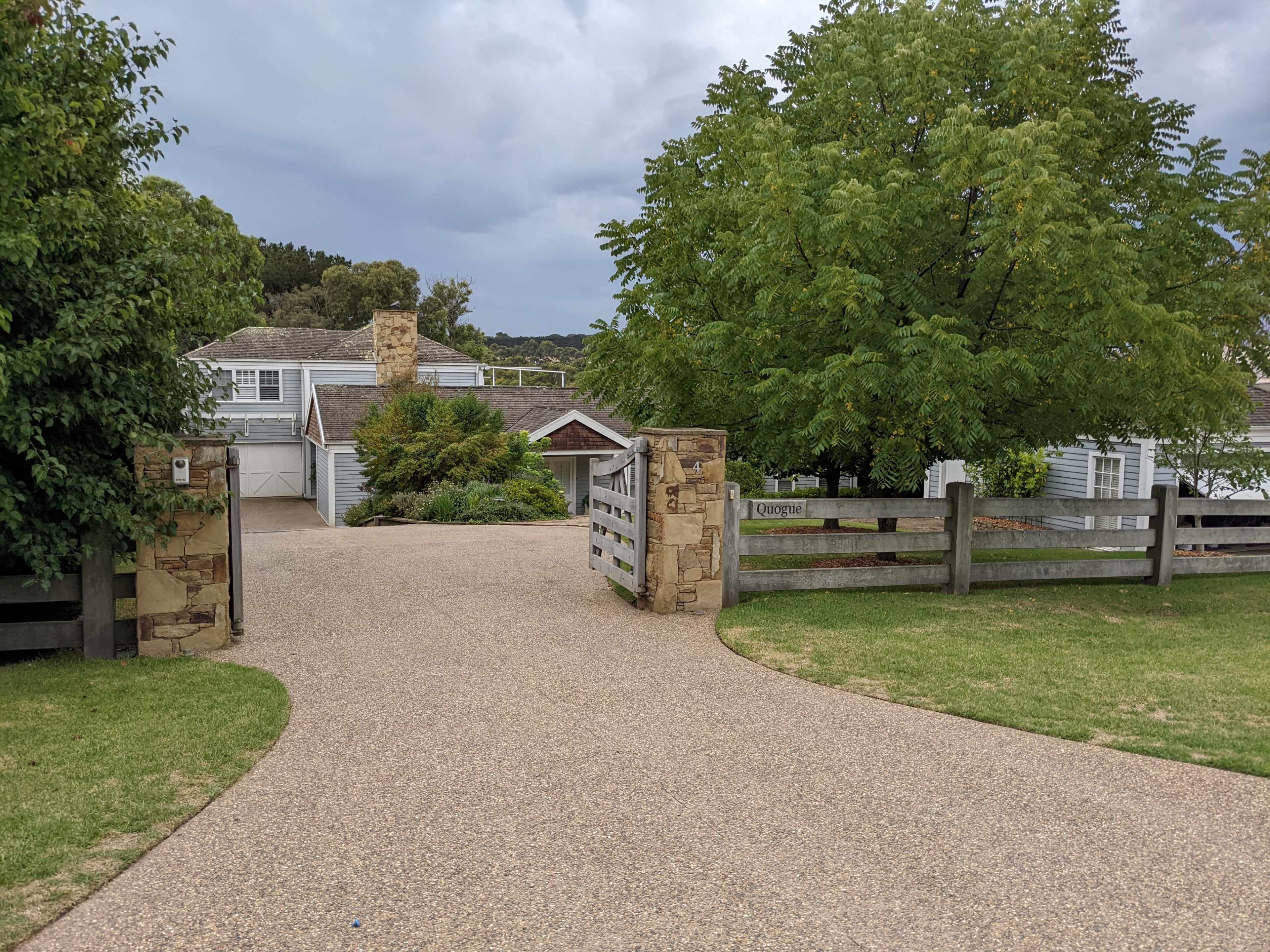 A gravel driveway leads to a house surrounded by greenery and trees, with a stone gate marking the entrance.